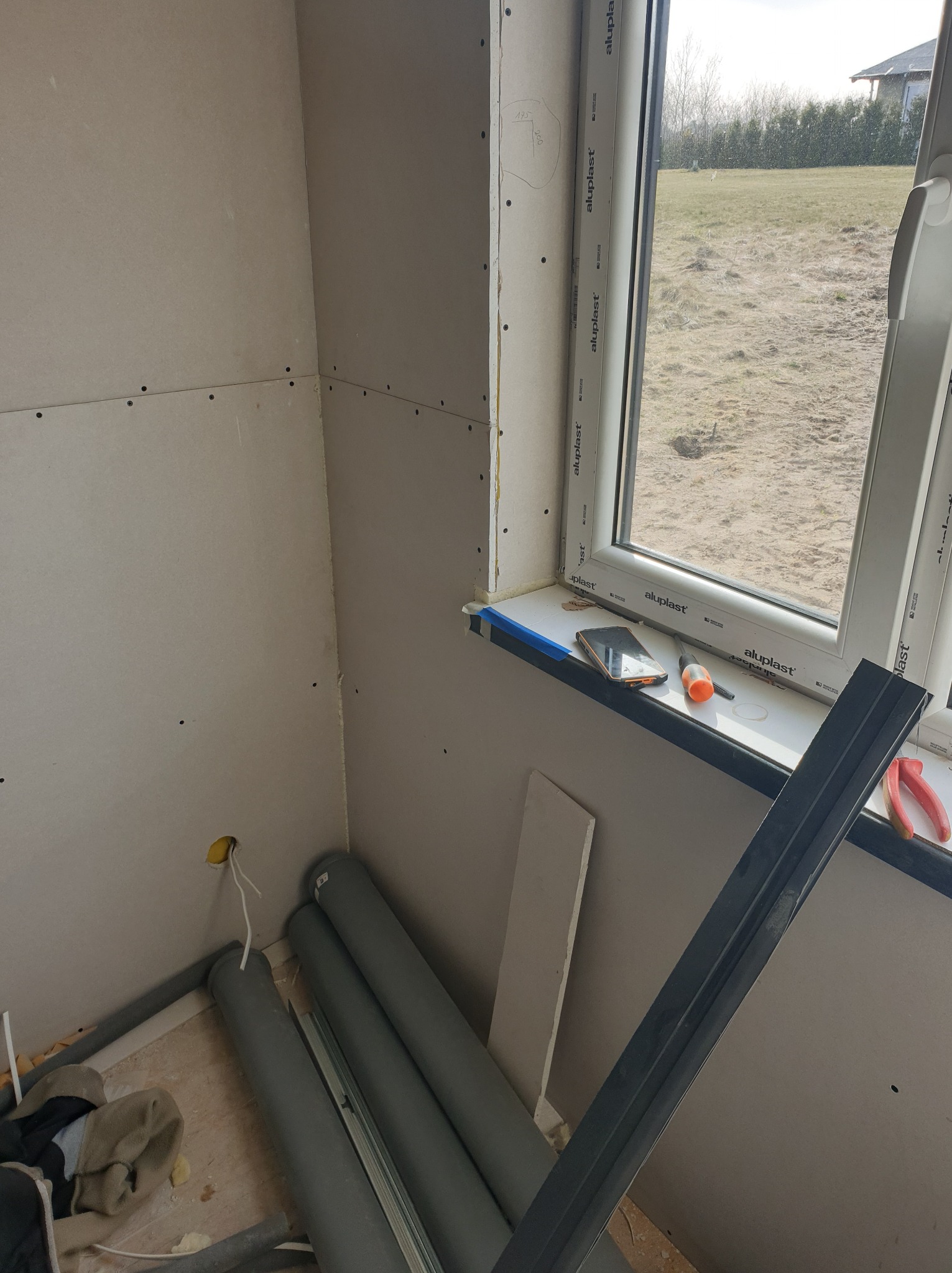 Wnętrze w trakcie prac wykończeniowych: ściany obłożone płytami kartonowo-gipsowymi, widoczne profile, przewody elektryczne wystające ze ściany oraz narzędzia na parapecie okiennym.