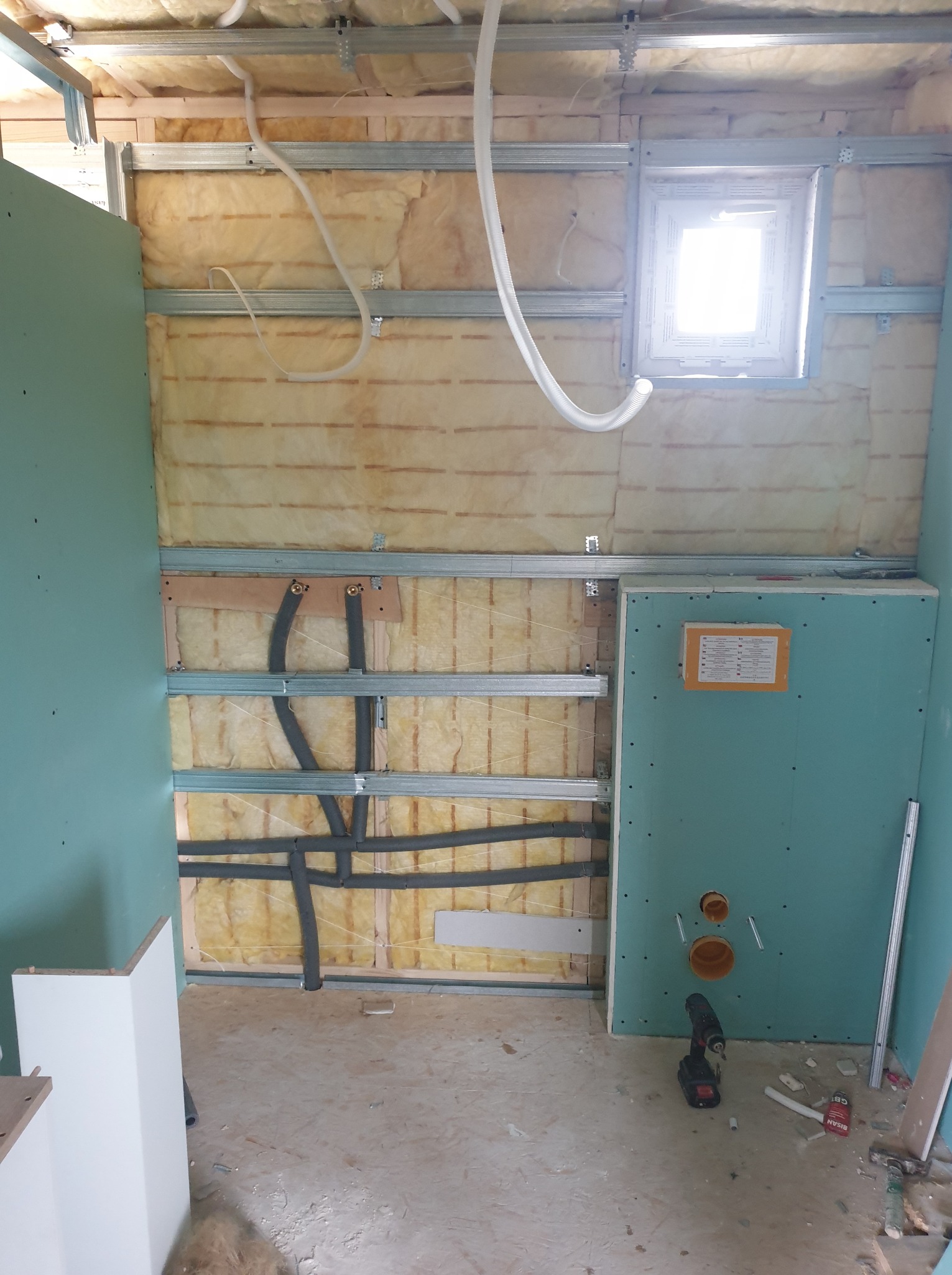 Wnętrze łazienki w trakcie remontu: widoczne ocieplenie ścian wełną mineralną, rury instalacji wodnej, częściowo zamontowane płyty kartonowo-gipsowe, okno z widocznym światłem dziennym, narzędzia...