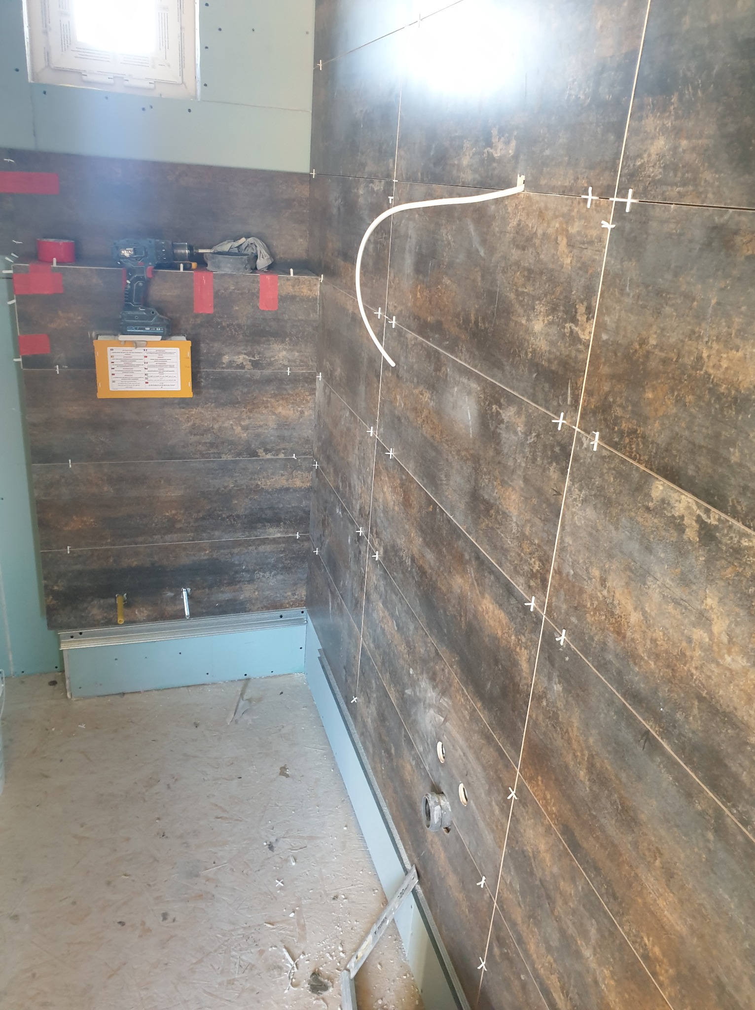 Układanie ciemnych płytek imitujących drewno w łazience, widoczne krzyżyki dystansowe, poziomica, wiertarka akumulatorowa i instrukcja obsługi na ścianie z płyt gipsowo-kartonowych.