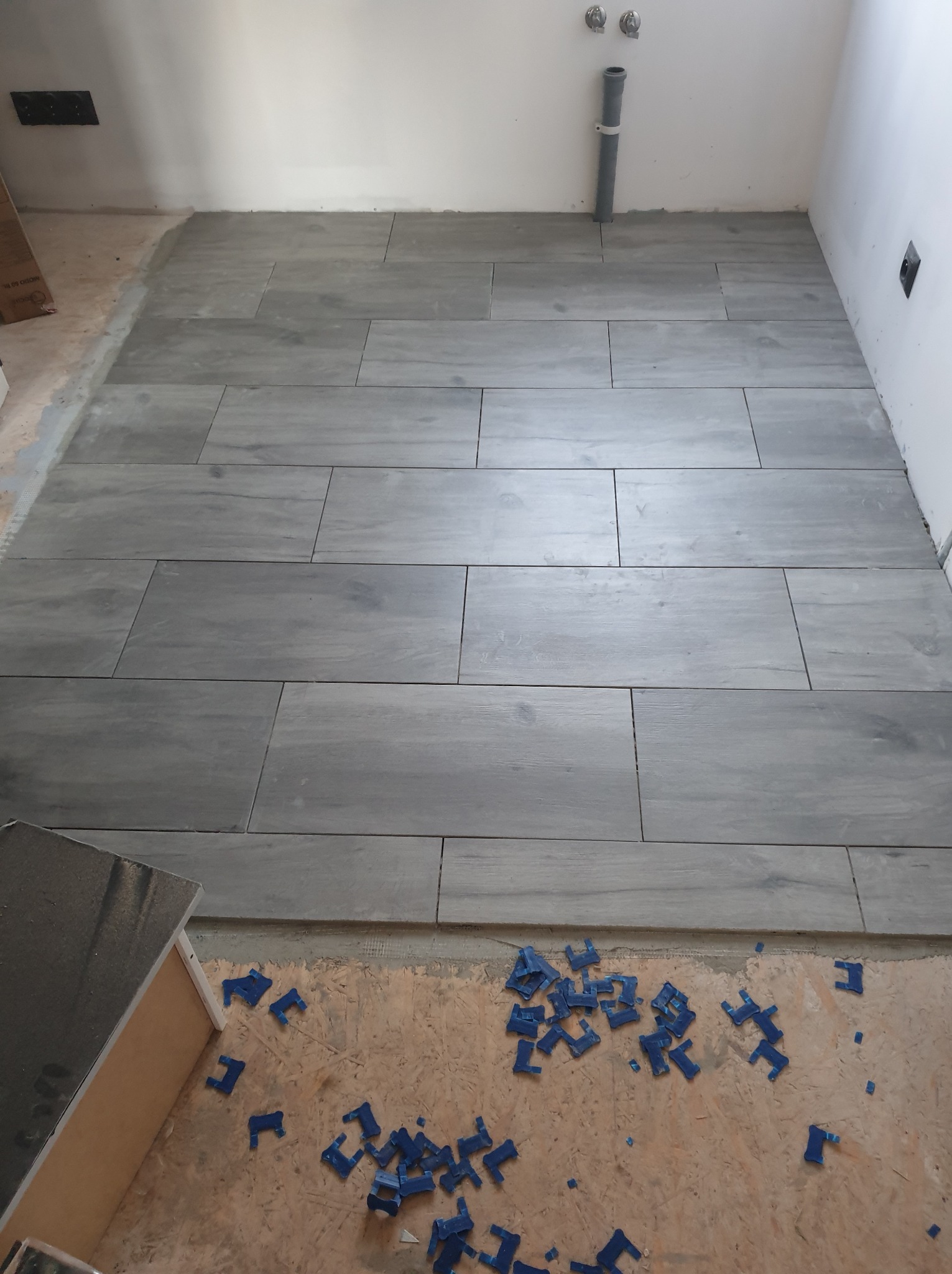 Układanie szarych płytek podłogowych imitujących drewno w pomieszczeniu, widoczne niebieskie klipsy do poziomowania płytek i rura kanalizacyjna przy ścianie.