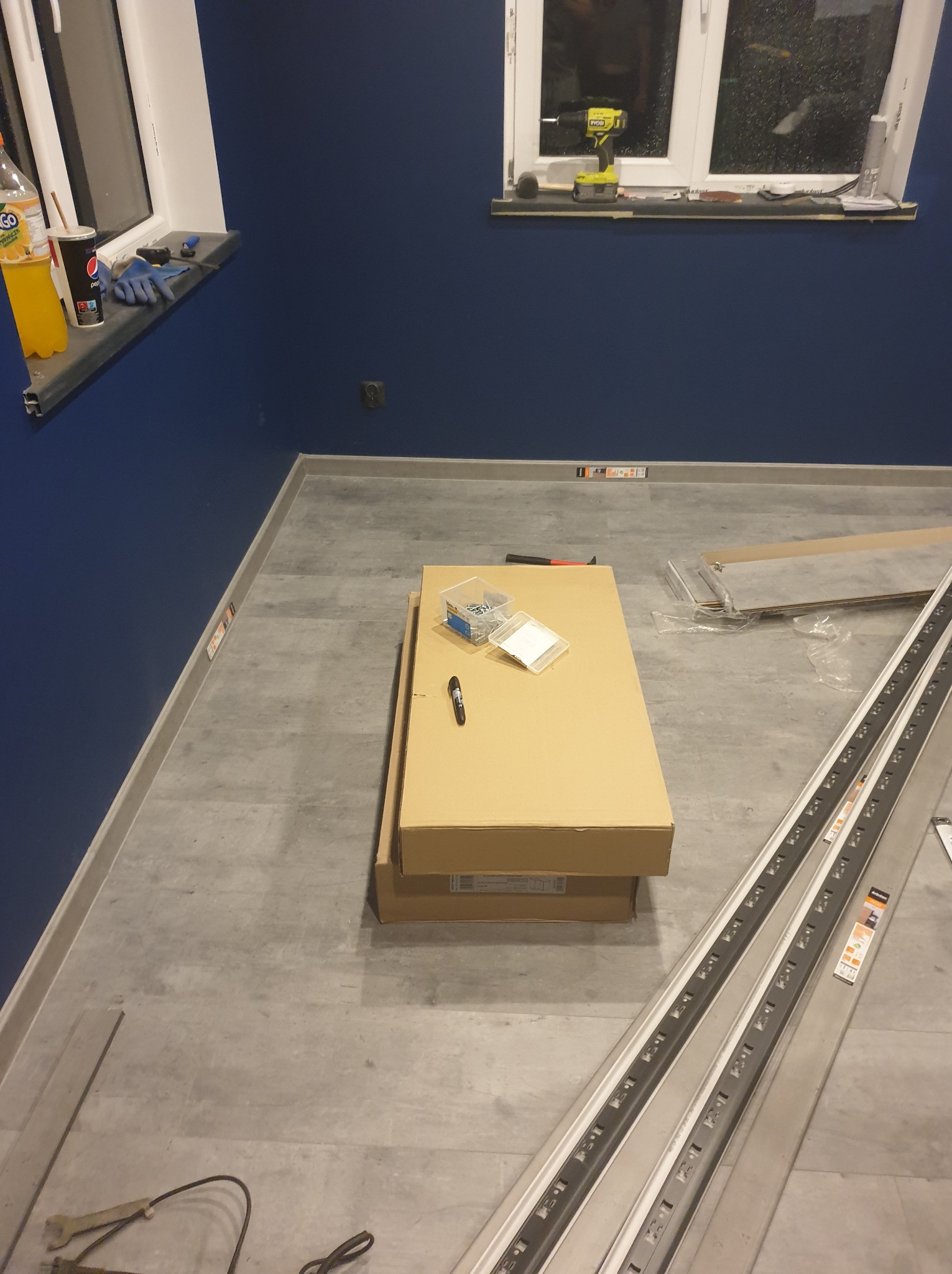 Przygotowania do montażu podłogi: panele, listwy przypodłogowe LED w opakowaniach, skrzynka z narzędziami i napoje na parapecie w pokoju z granatowymi ścianami.