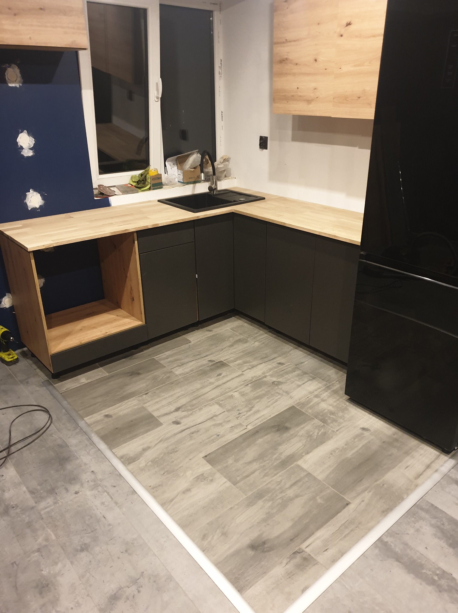 Częściowo zmontowana kuchnia z drewnianym blatem i ciemnoszarymi szafkami, widoczne niedokończone ściany i podłoga z płytek imitujących drewno, w tle okno i czarna lodówka.