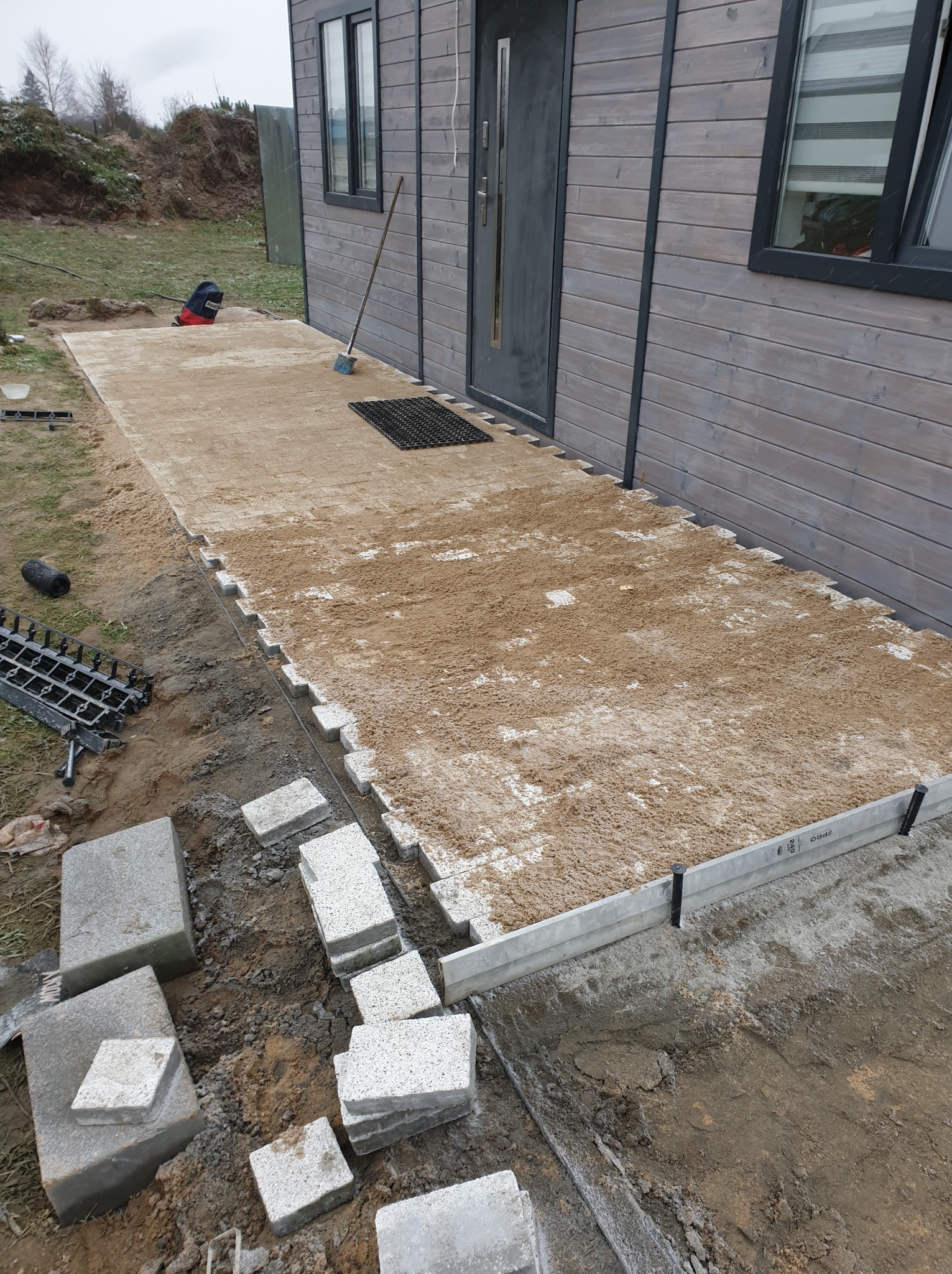 Układanie kostki brukowej przed domem z drewnianą elewacją, widoczne narzędzia i materiały budowlane, etap przygotowania podłoża z podsypką piaskową.