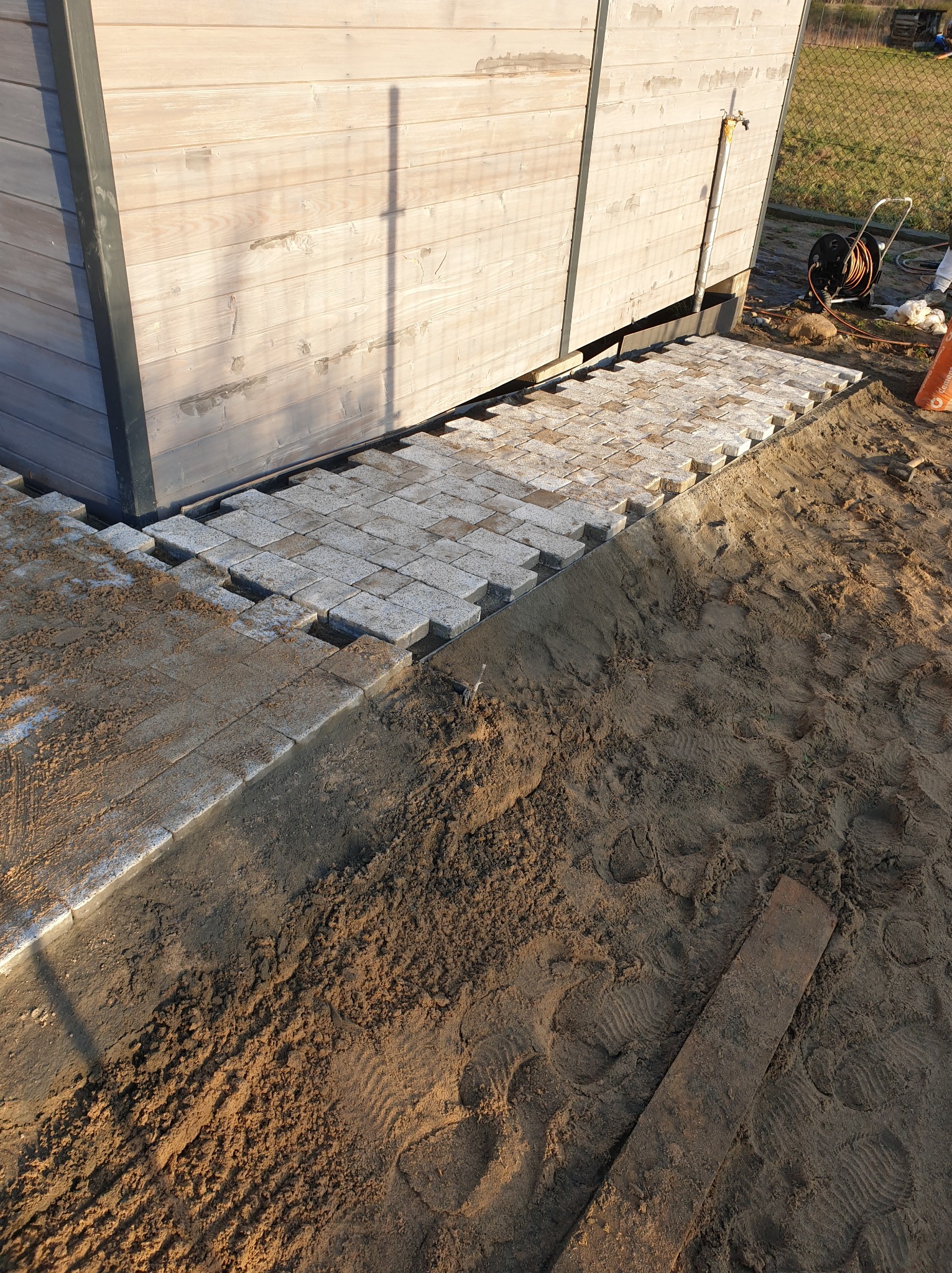 Układanie szarej kostki brukowej wokół drewnianej konstrukcji, widoczny piasek i ziemia, etap prac.