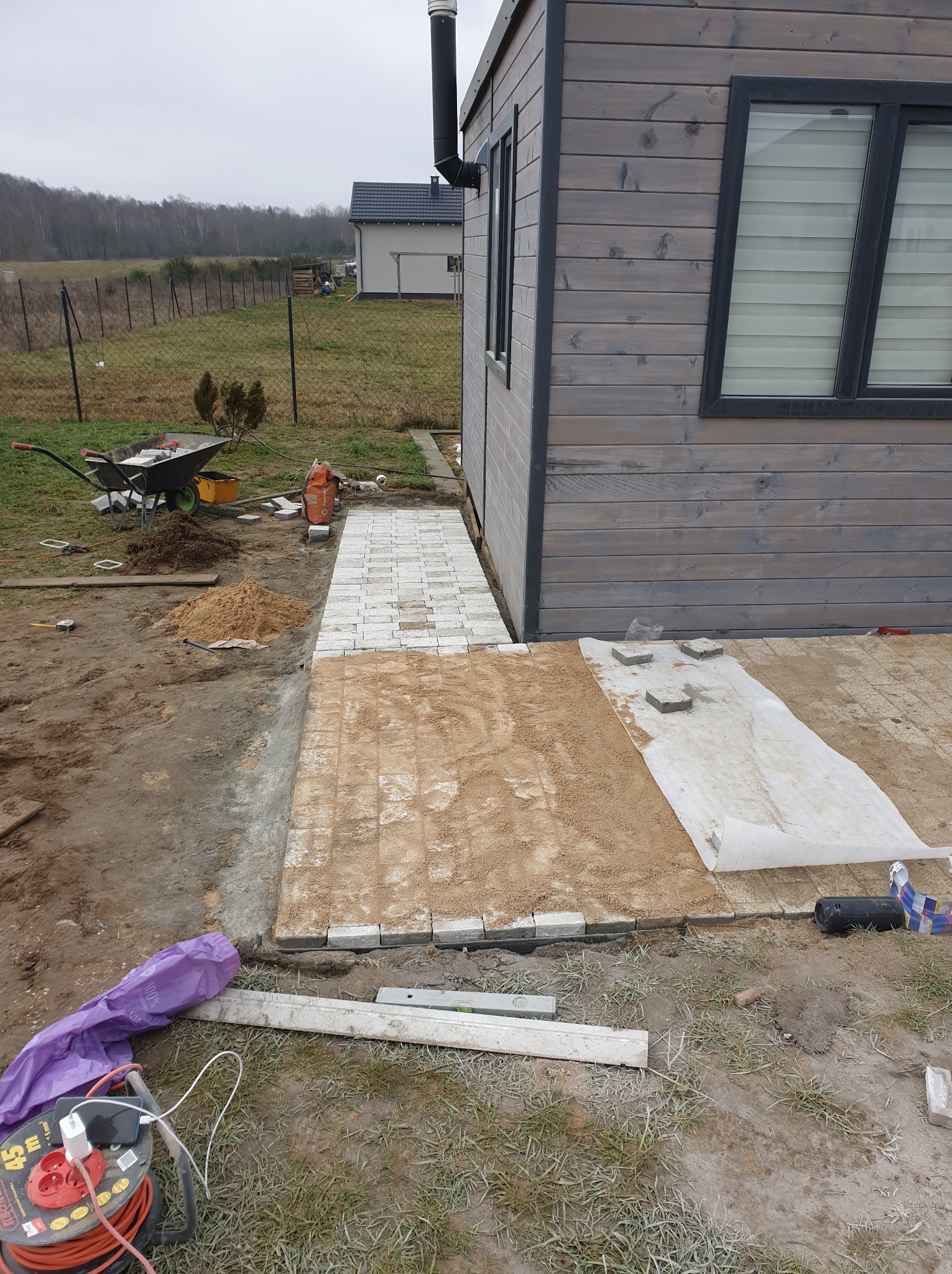 Układanie jasnoszarej kostki brukowej na podsypce piaskowej obok drewnianego domku letniskowego; widoczna taczka z kostką, poziomica i zwinięty przedłużacz elektryczny.