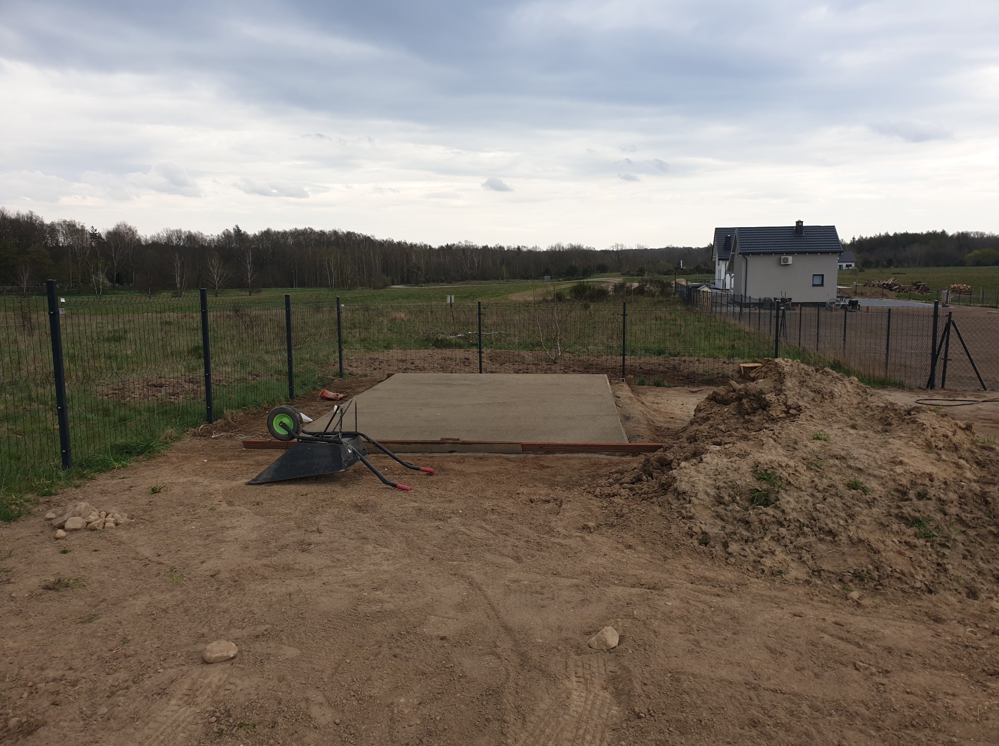 Betonowa wylewka fundamentowa w trakcie prac, widoczna taczka i sterta ziemi, dom w tle, pochmurne niebo.