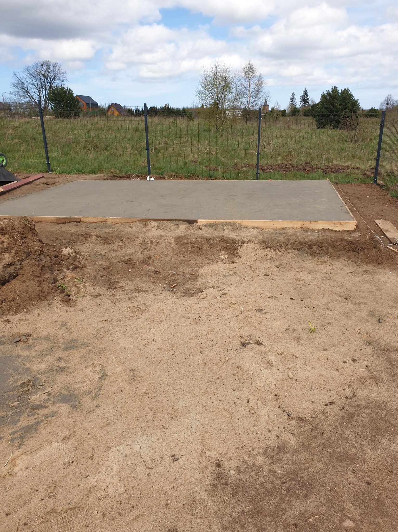 Wylana betonowa płyta fundamentowa z drewnianym szalunkiem na piaszczystym terenie działki, widoczna siatka ogrodzeniowa i zieleń w tle.