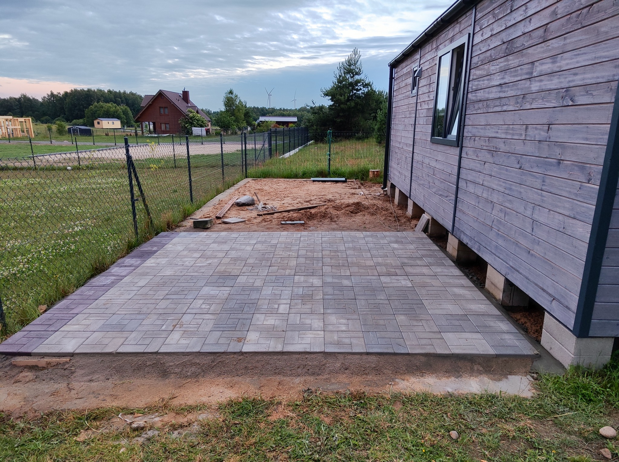 Ułożona szara kostka brukowa tworząca taras przy drewnianym domu na tle wiejskiego krajobrazu z wiatrakami.