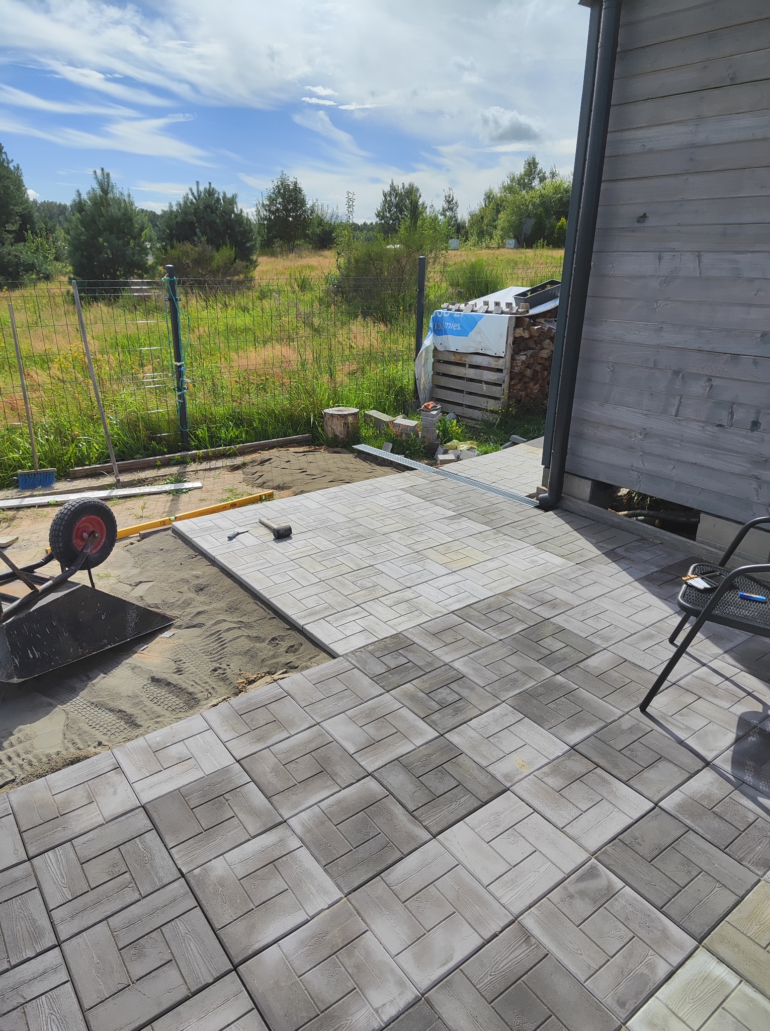 Układanie szarej kostki brukowej o wzorze drewna na podsypce piaskowej przy drewnianym budynku, widoczny wózek z narzędziami i poziomica.