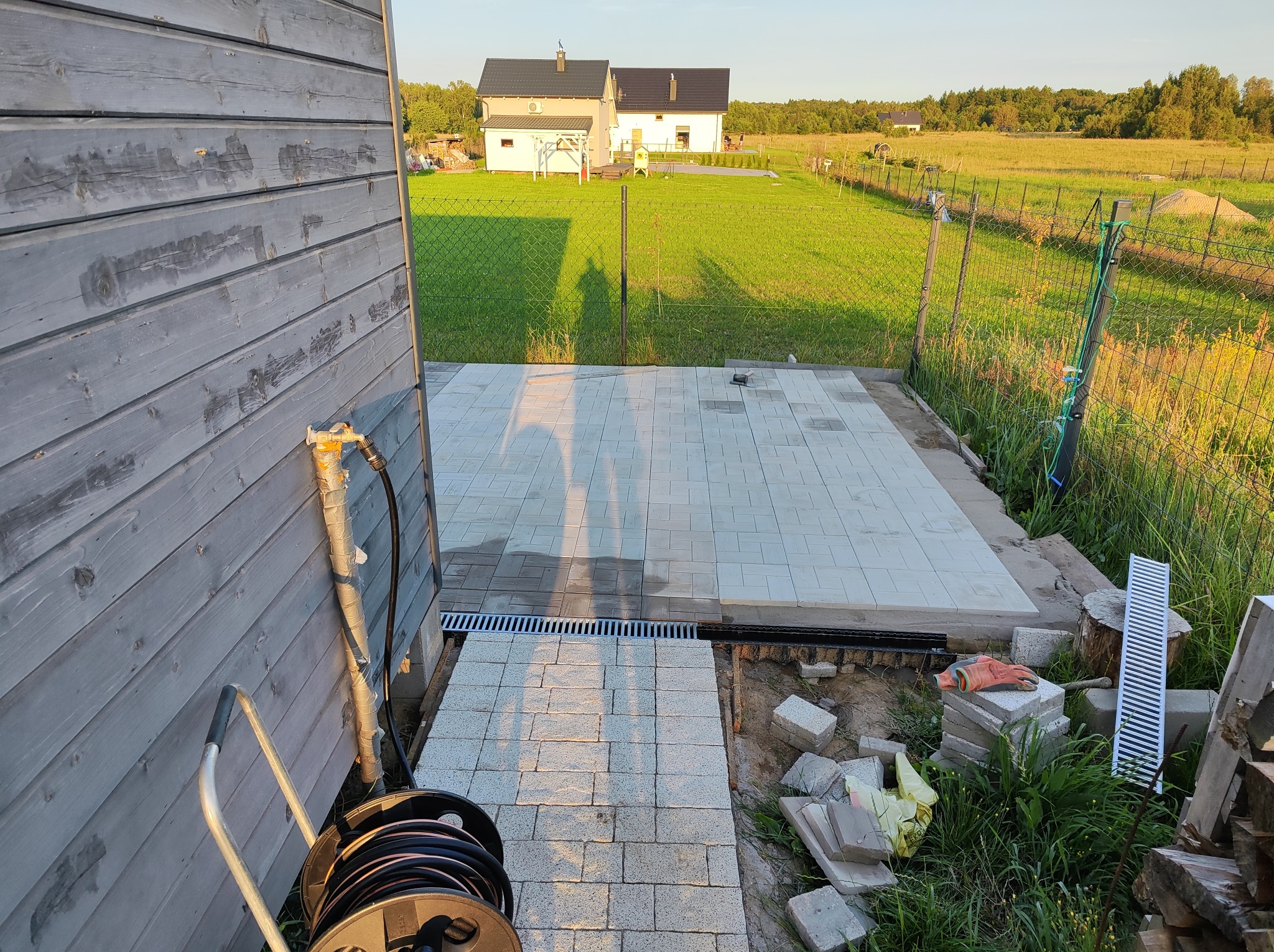 Układanie jasnoszarej kostki brukowej na tarasie w ogrodzie, widoczny fragment drewnianej elewacji domu, trawnik i drugi dom w tle, narzędzia pracy i zapas kostki obok.
