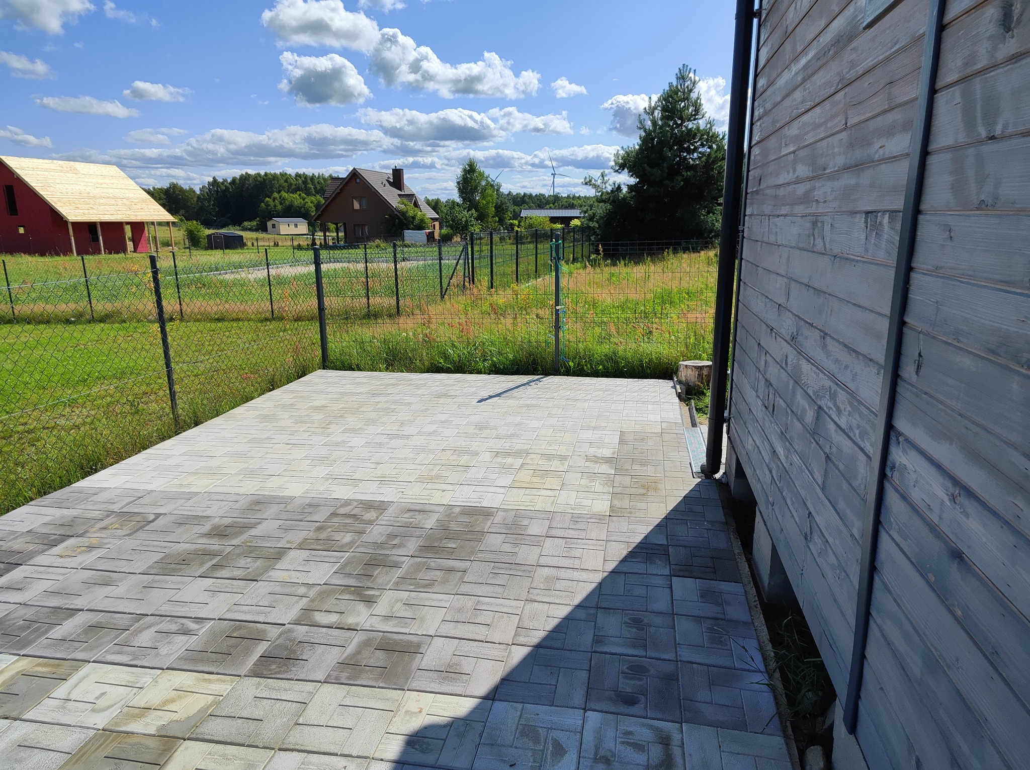 Nowo ułożony, szary chodnik z kostki brukowej, przylegający do drewnianego domu, z widokiem na okoliczne budynki i las w tle, w słoneczny dzień.