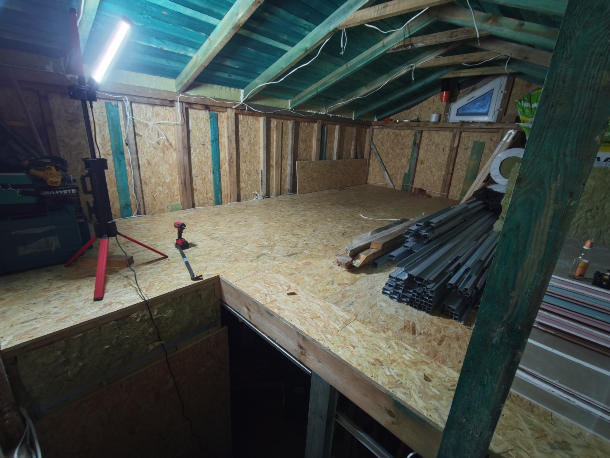 Poddasze w trakcie adaptacji: drewniana konstrukcja, płyty OSB na podłodze i ścianach, narzędzia pracy, profile metalowe. Widoczne oświetlenie robocze.