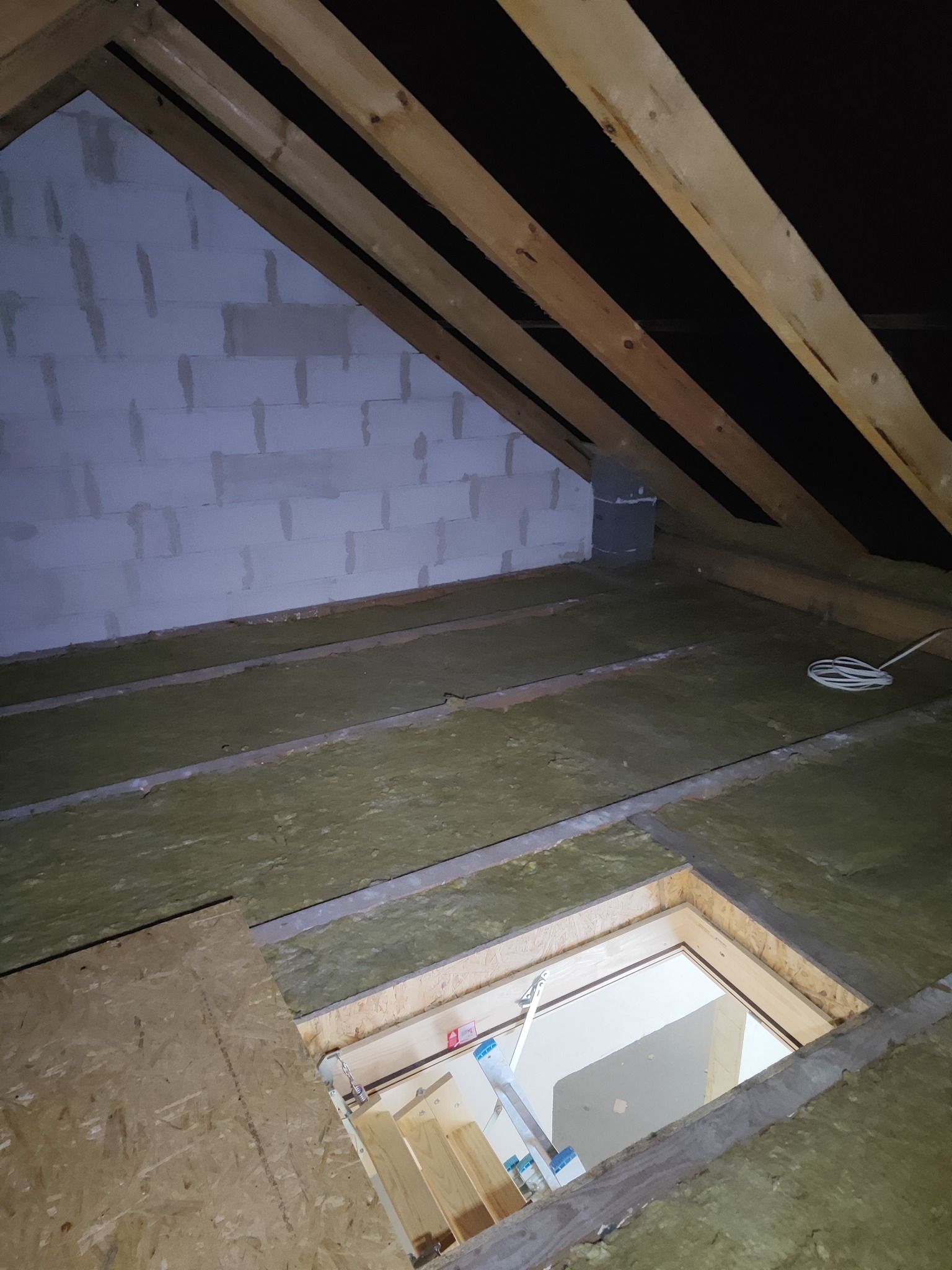 Ocieplone poddasze z widoczną konstrukcją drewnianą, wełną mineralną i otworem włazowym z drabinką. Ściana z bloczków betonu komórkowego.