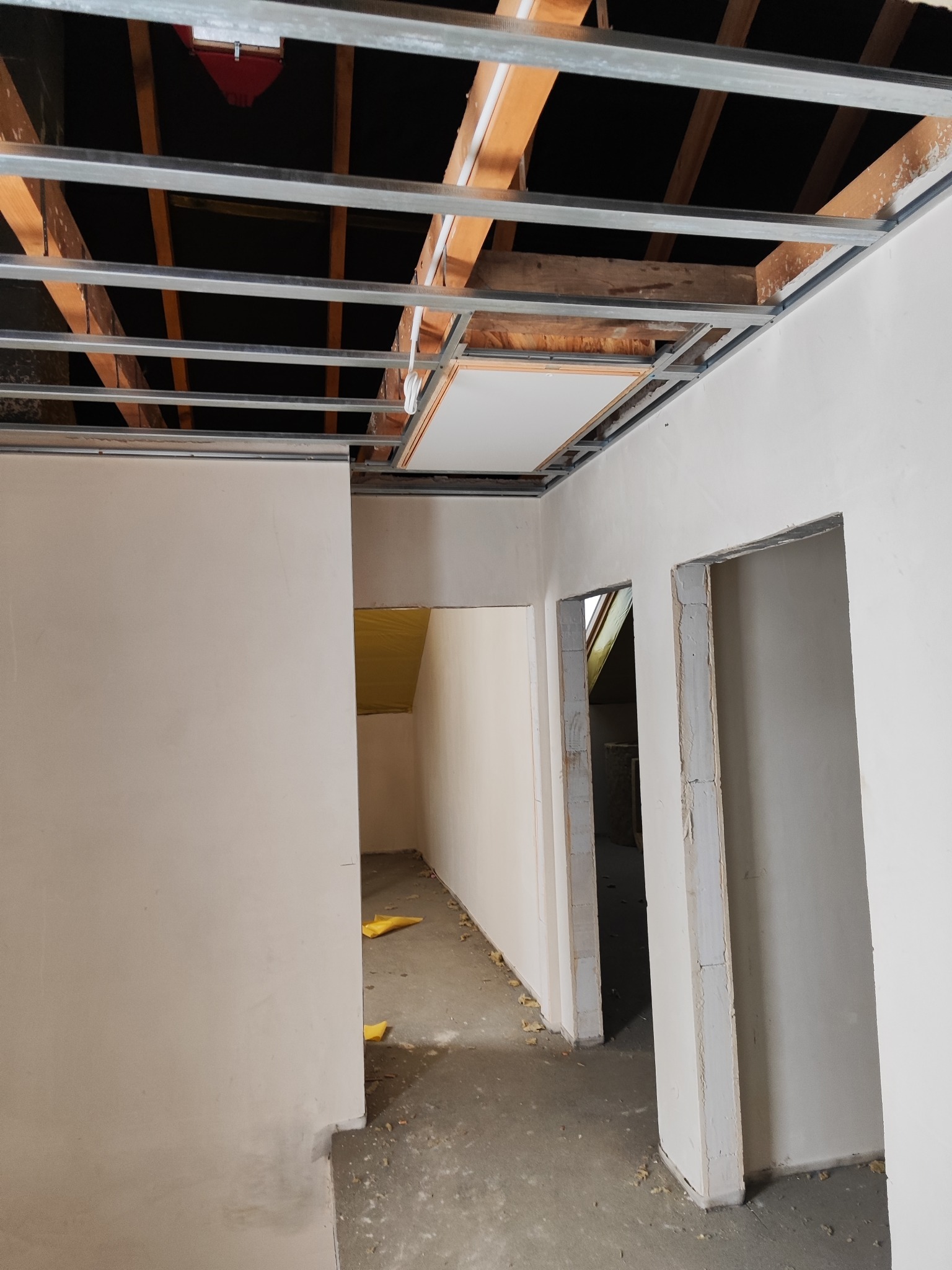 Wnętrze w trakcie prac wykończeniowych: metalowa konstrukcja sufitu podwieszanego, ściany po gładziowaniu, przygotowane otwory drzwiowe. Widok z dołu.