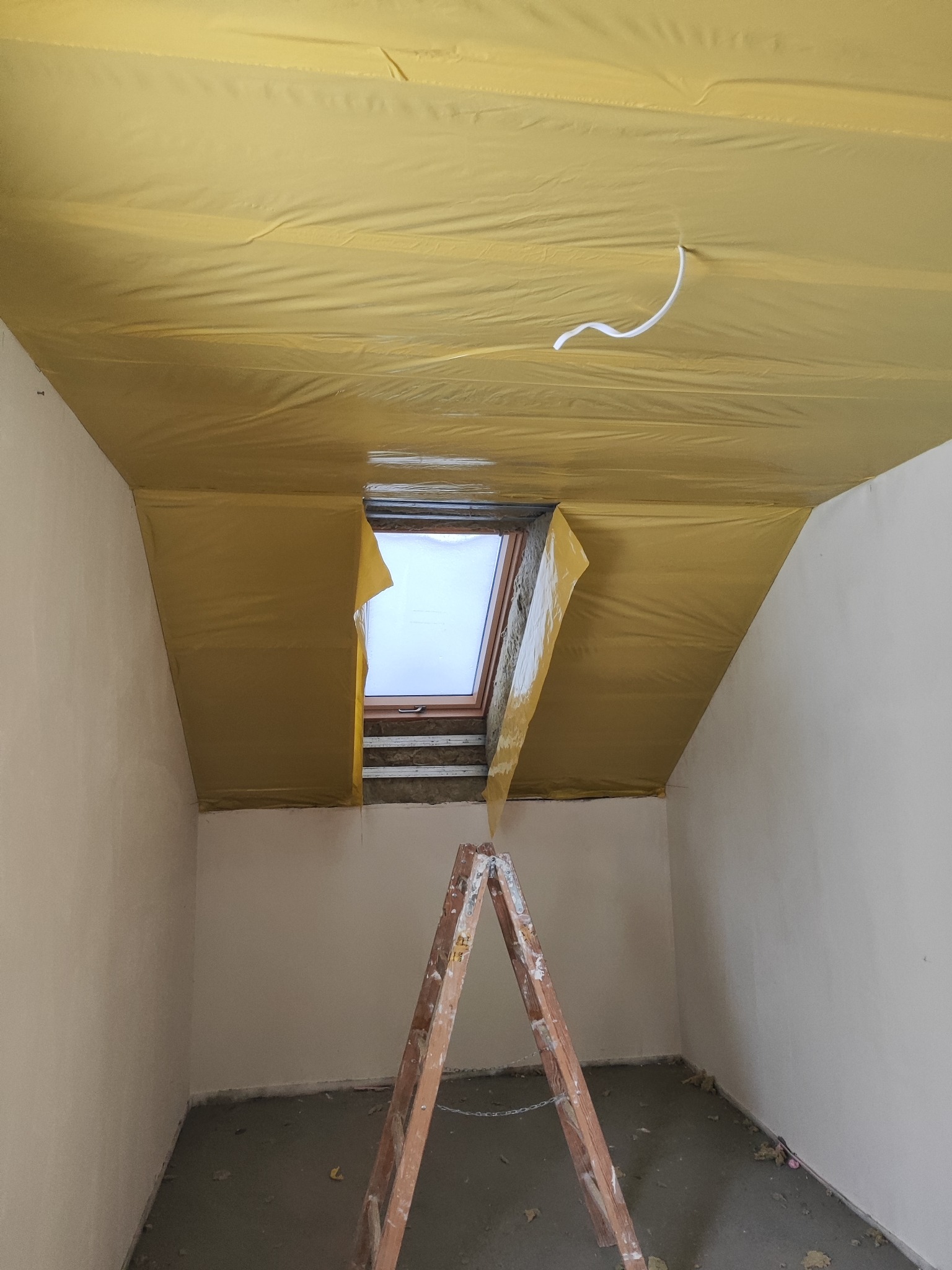 Poddasze z oknem dachowym w trakcie prac wykończeniowych. Widoczna folia paroizolacyjna na suficie i ściany przygotowane do dalszych prac. Stoi drabina.