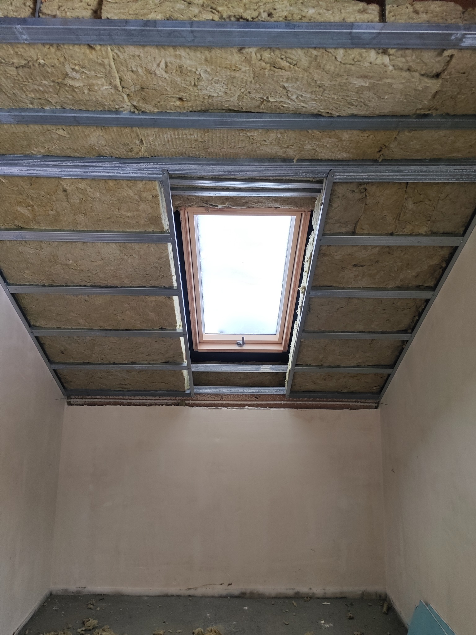 Montaż izolacji z wełny mineralnej w metalowym stelażu sufitu podwieszanego z oknem dachowym. Widok od dołu na sufit w trakcie prac wykończeniowych.