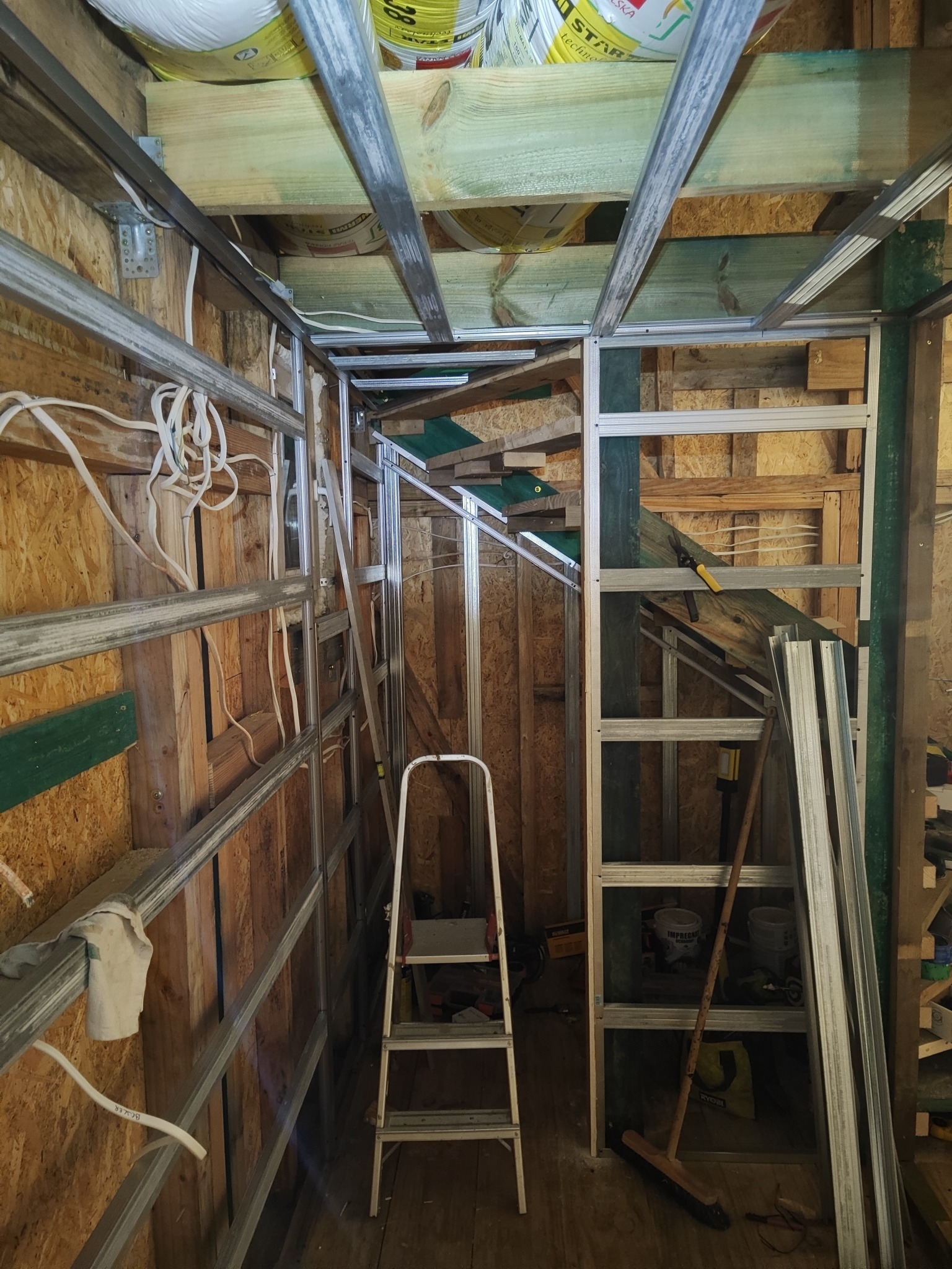 Szkielet konstrukcji z profili metalowych na drewnianej ścianie, widoczne przewody elektryczne i drabina. Przygotowanie do montażu płyt gipsowo-kartonowych.