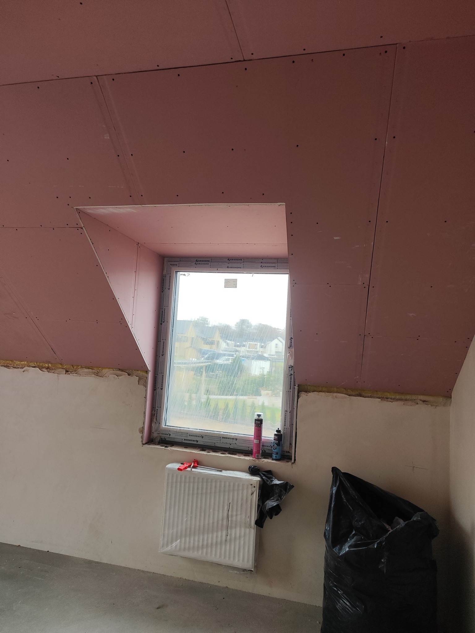 Montaż płyt karton-gips na poddaszu z oknem dachowym. Widoczne łączenia płyt i wkręty. Częściowo wykończona ściana, grzejnik i worek na śmieci przy oknie.
