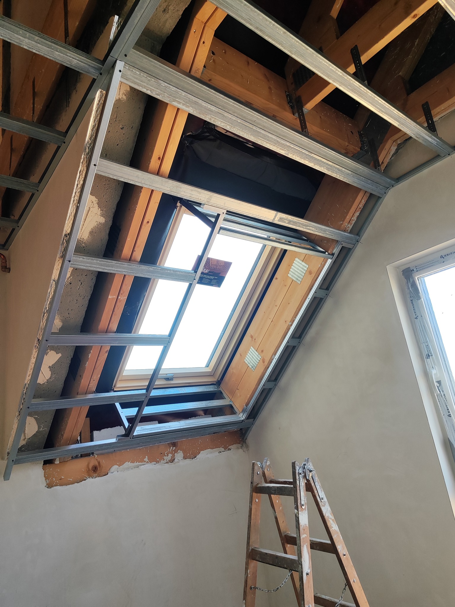 Montaż sufitu podwieszanego z płyt karton-gips wokół okna dachowego. Widoczne metalowe profile i drewniana konstrukcja dachu. Drabina oparta o ścianę.