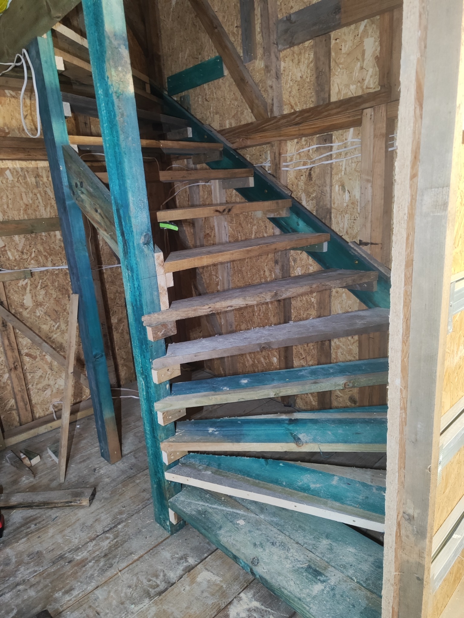 Drewniane schody w trakcie budowy, z pomalowanymi na turkusowo elementami konstrukcyjnymi. Widoczne surowe deski i fragmenty ścian z płyt OSB. Ujęcie z dołu, podkreślające konstrukcję.