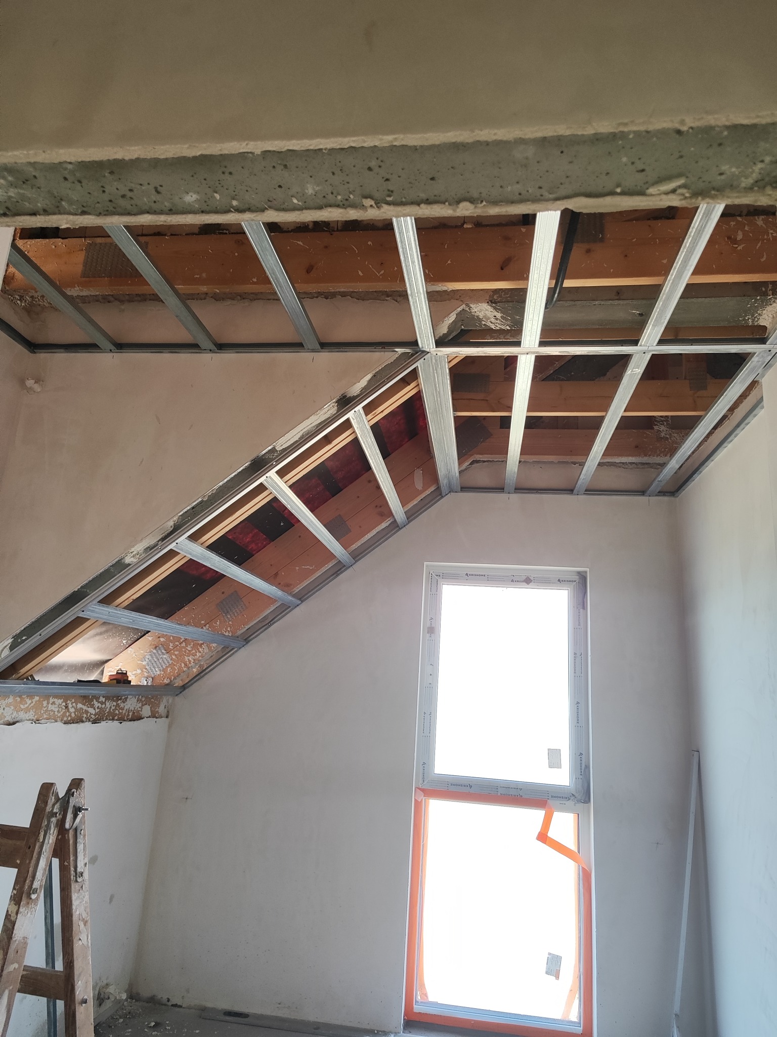 Skelet sufitu podwieszanego z profili metalowych w pokoju na poddaszu, widoczne drewniane elementy konstrukcyjne. Okno z pomarańczową folią ochronną.