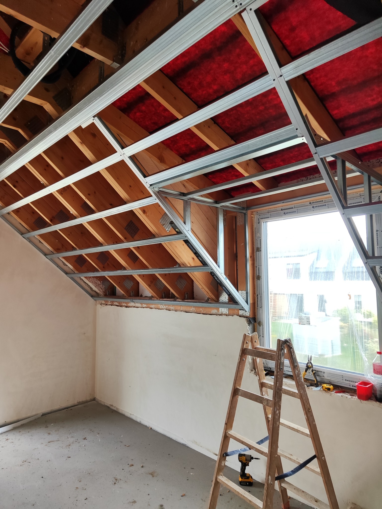 Montaż sufitu podwieszanego z płyt karton-gips na poddaszu w Salino. Widoczne profile, drewniana konstrukcja dachu i okno. Prace wykończeniowe w trakcie.