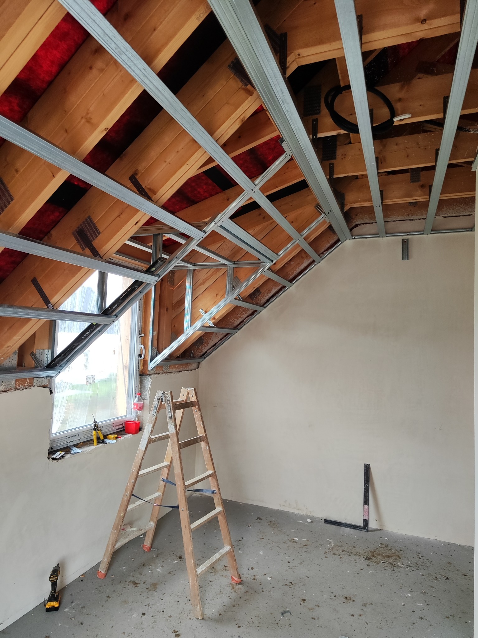 Szkielet sufitu podwieszanego z profili stalowych w poddaszu, widoczne ocieplenie, drabina i narzędzia. Ściany przygotowane pod gładź.