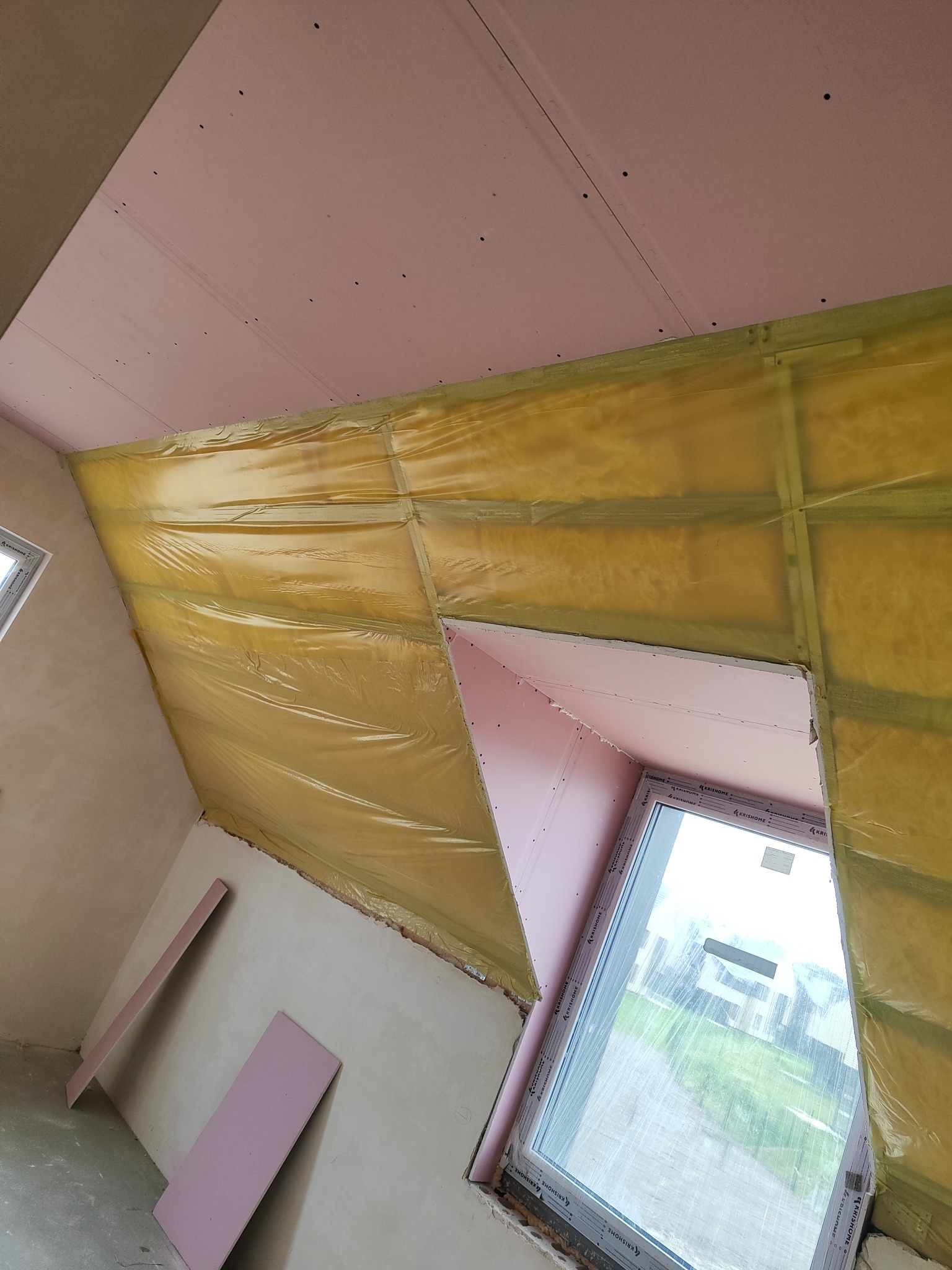 Montaż płyty karton-gips na poddaszu z oknem dachowym. Widoczna izolacja folią paroizolacyjną. Fragment ściany z surowym tynkiem. Dwie płyty leżą na podłodze.