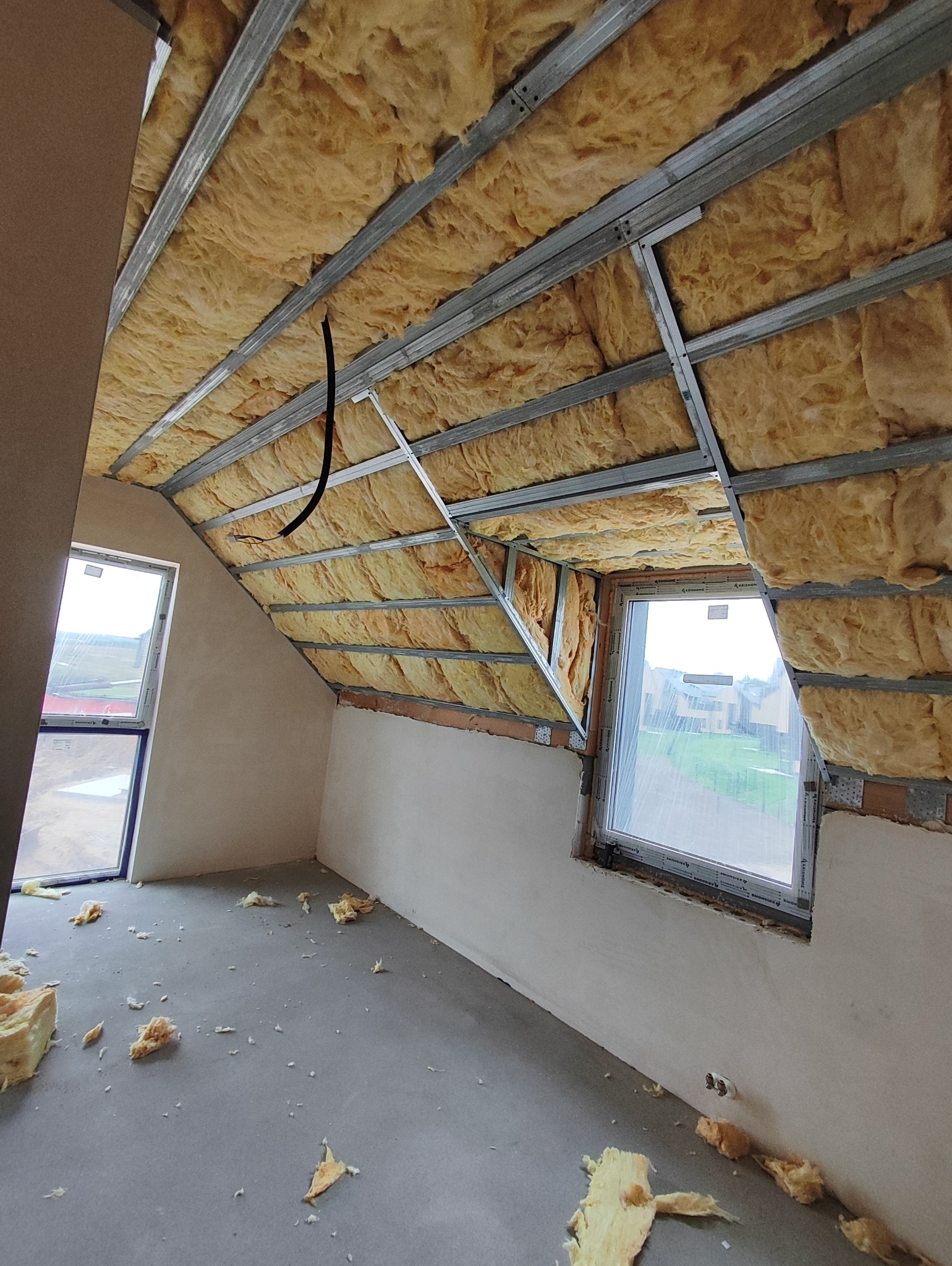 Poddasze w trakcie izolacji: wełna mineralna między metalowymi profilami, okna dachowe, surowy beton na podłodze z resztkami izolacji. Etap prac wykończeniowych.