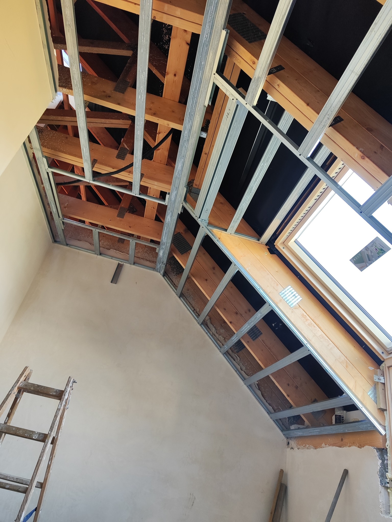 Montaż stelażu z profili stalowych pod płytę gipsową na poddaszu z oknem dachowym. Widoczna drewniana konstrukcja dachu i drabina oparta o ścianę.