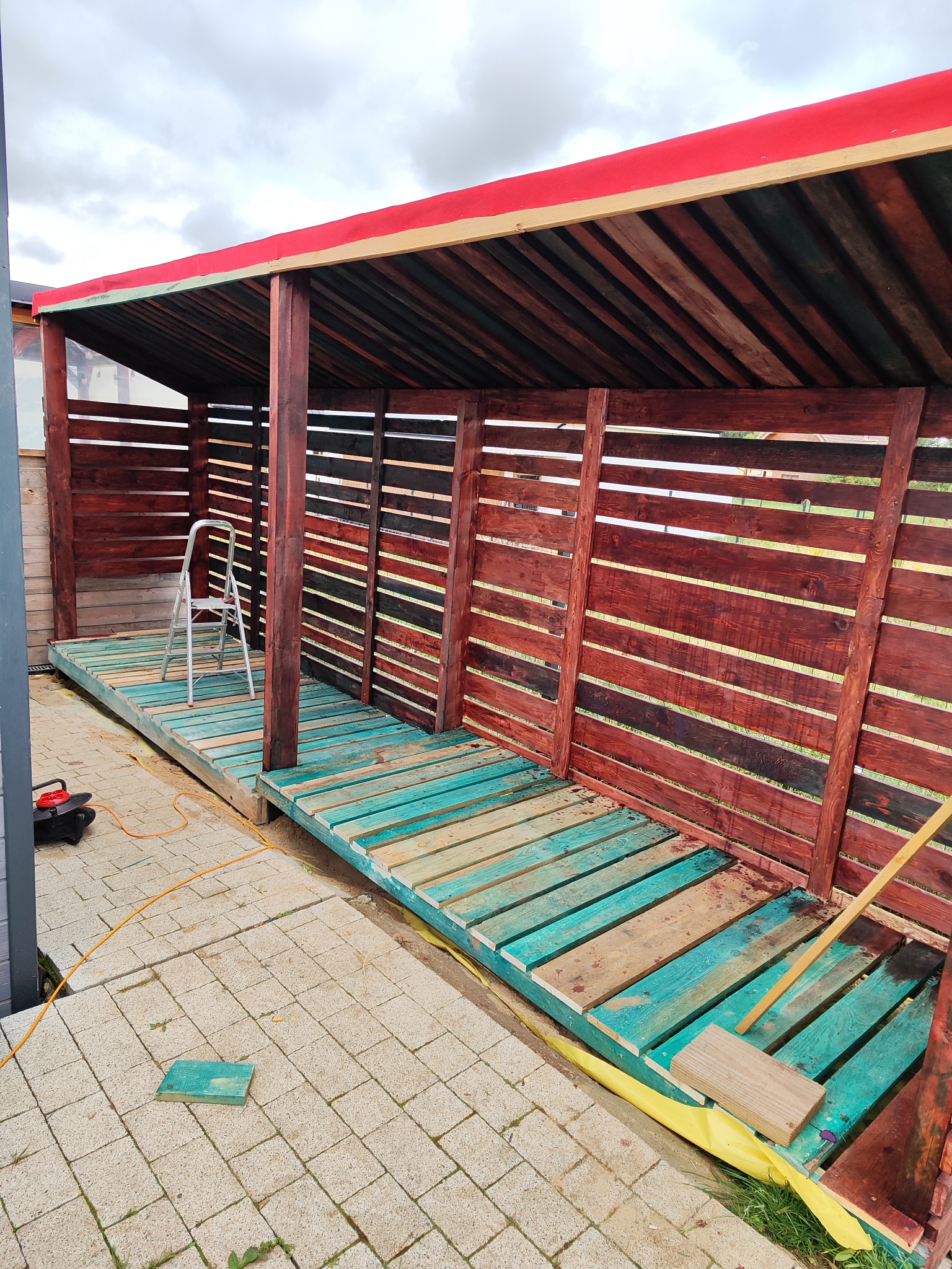 Drewniana konstrukcja z czerwonym dachem w trakcie budowy. Widoczne deski pomalowane na turkusowo i brązowo, drabina i narzędzia. Prace wykończeniowe w ogrodzie.
