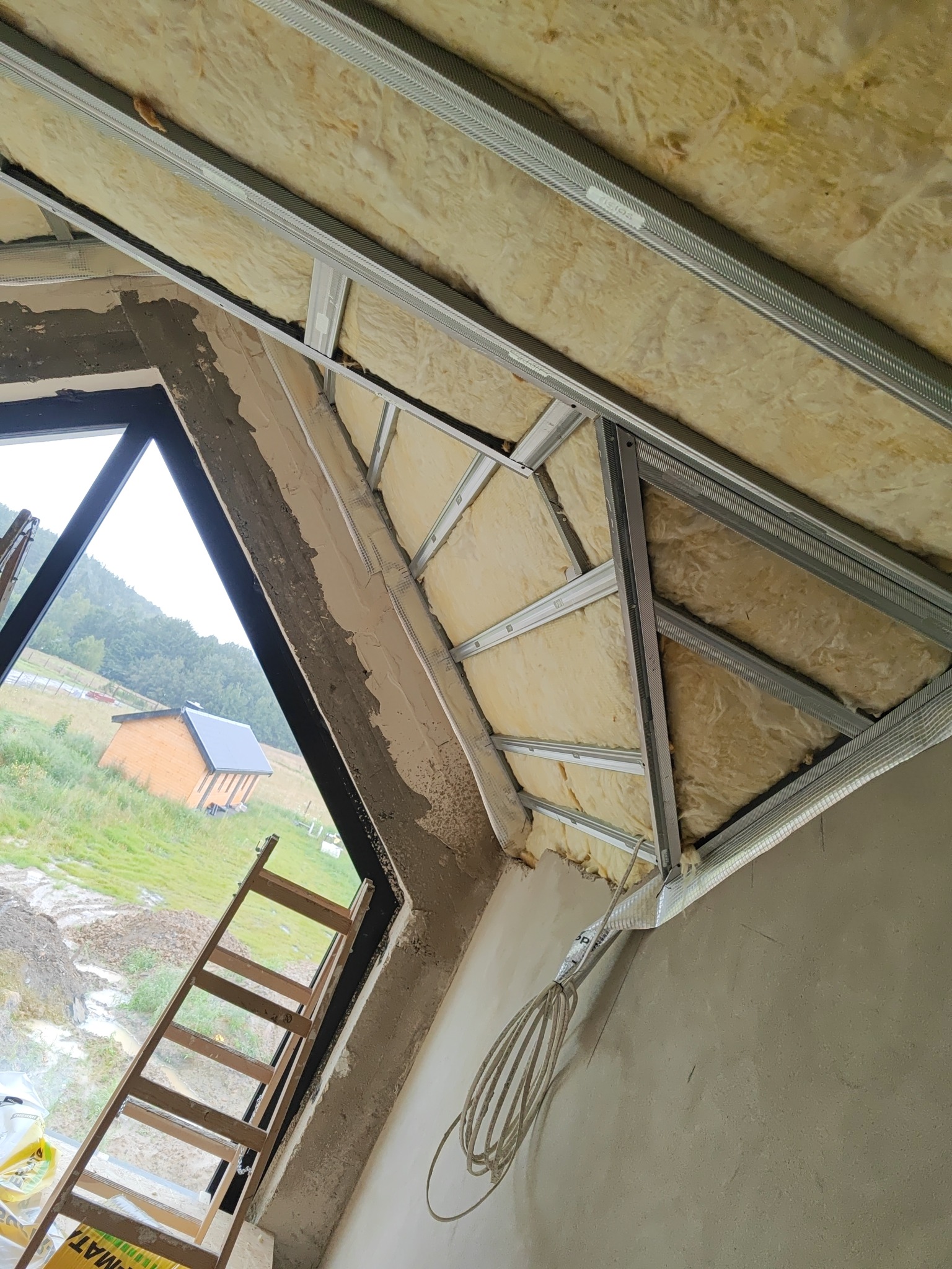 Skośny sufit poddasza w trakcie izolacji wełną mineralną, widoczna metalowa konstrukcja stelaża, okno z widokiem na krajobraz i drabina oparta o ścianę.