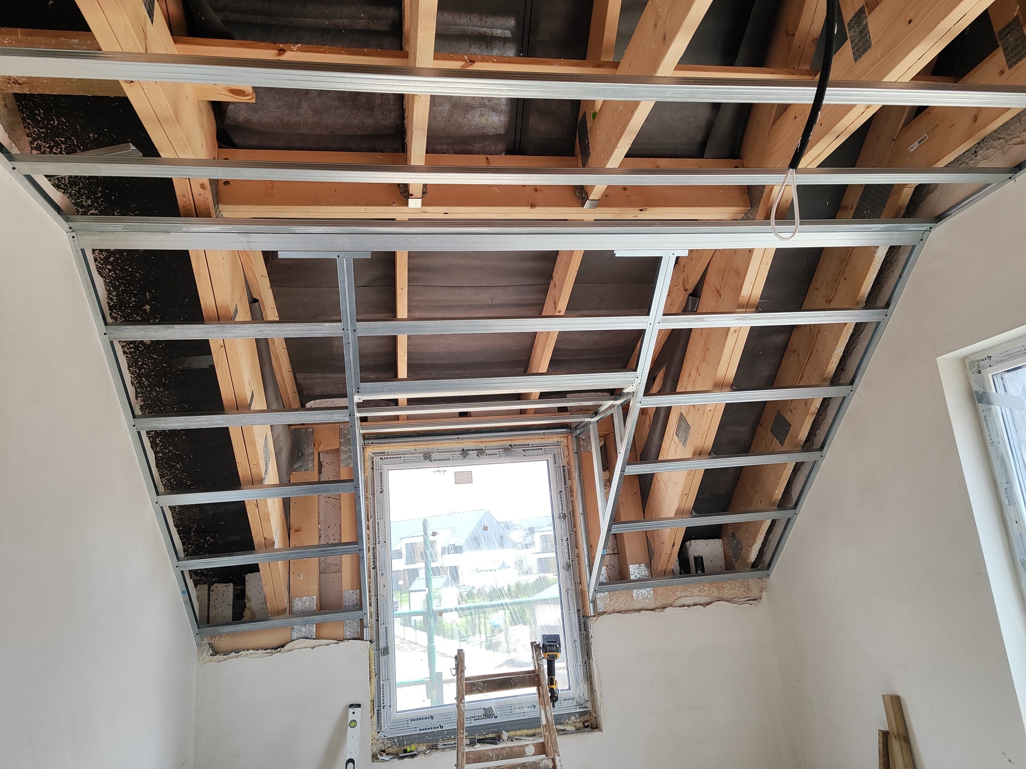 Montaż metalowej konstrukcji sufitu podwieszanego na poddaszu z drewnianą więźbą. Widoczne okno dachowe i drabina. Etap prac przed montażem płyt.