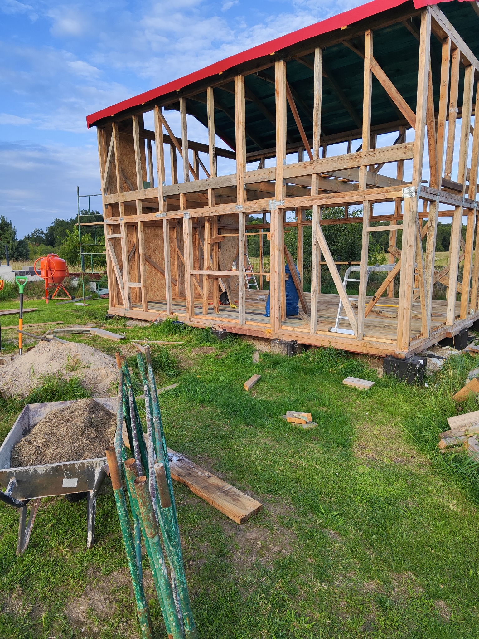 Szkielet drewnianej konstrukcji budynku z czerwonym dachem w trakcie budowy, widoczne narzędzia i betoniarka w tle, na trawiastym terenie.