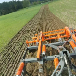 Pomarańczowo-szary agregat uprawowy w trakcie pracy na polu, widok z perspektywy operatora, świeżo spulchniona ziemia w rzędach, zielone pole w tle, drzewa na horyzoncie.