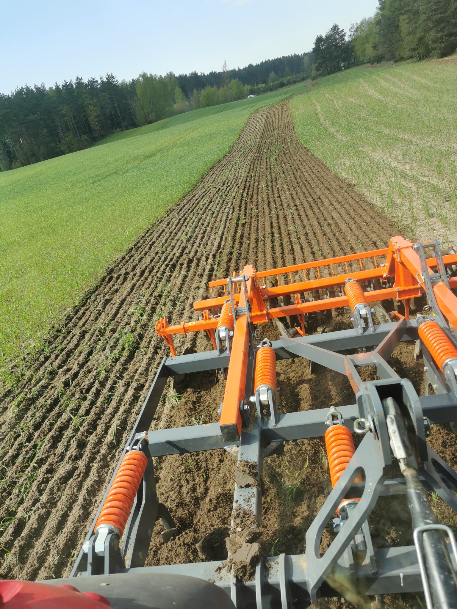 Pomarańczowo-szary agregat uprawowy w trakcie pracy na polu, widok z perspektywy operatora, świeżo spulchniona ziemia w rzędach, zielone pole w tle, drzewa na horyzoncie.