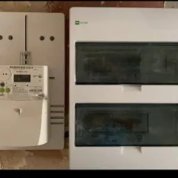 Nowy licznik energii elektrycznej GAMA 100 obok białej skrzynki bezpiecznikowej, zamontowane na ścianie z surową fakturą. Widoczne detale montażu i podłączenia.