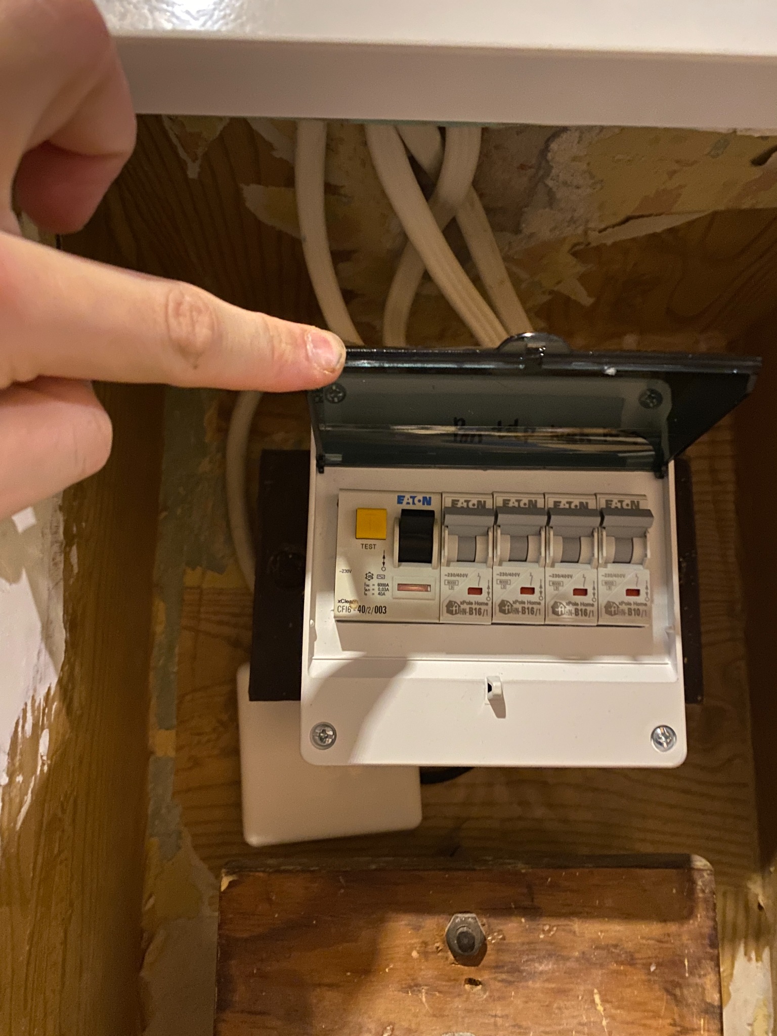 Otwarta skrzynka bezpiecznikowa marki Eaton z białymi przewodami w tle. Palec wskazujący na górną krawędź skrzynki. Widoczne ślady remontu w otoczeniu.
