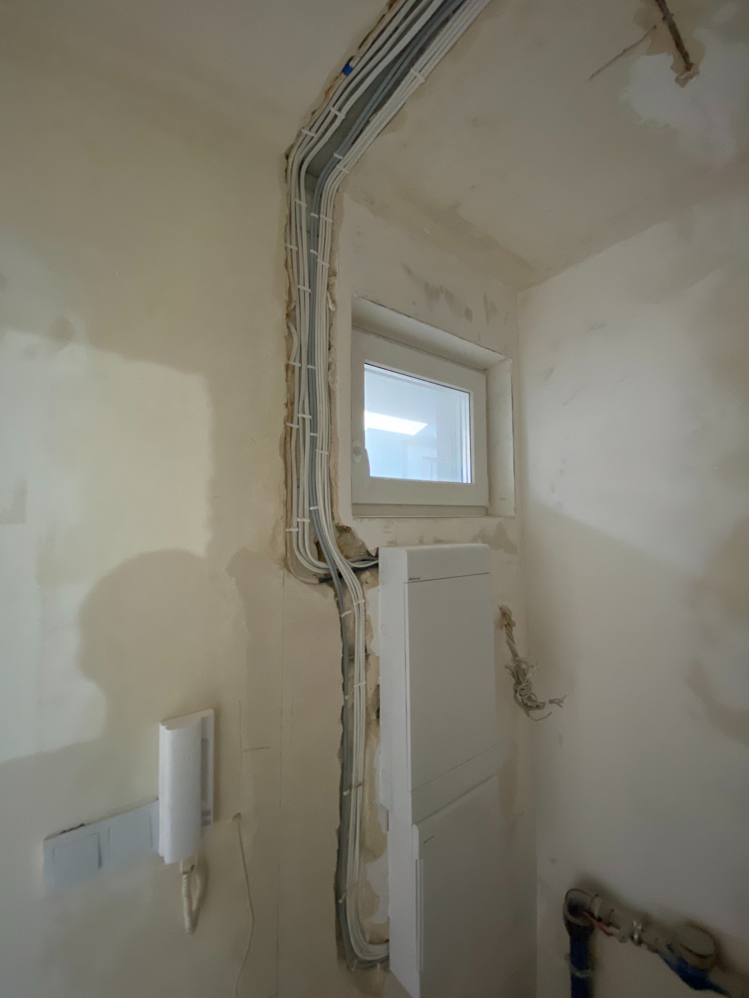 Białe kable elektryczne prowadzone wzdłuż ściany w nowym budynku, widoczne puszki i instalacje podtynkowe przed wykończeniem, okno w tle.