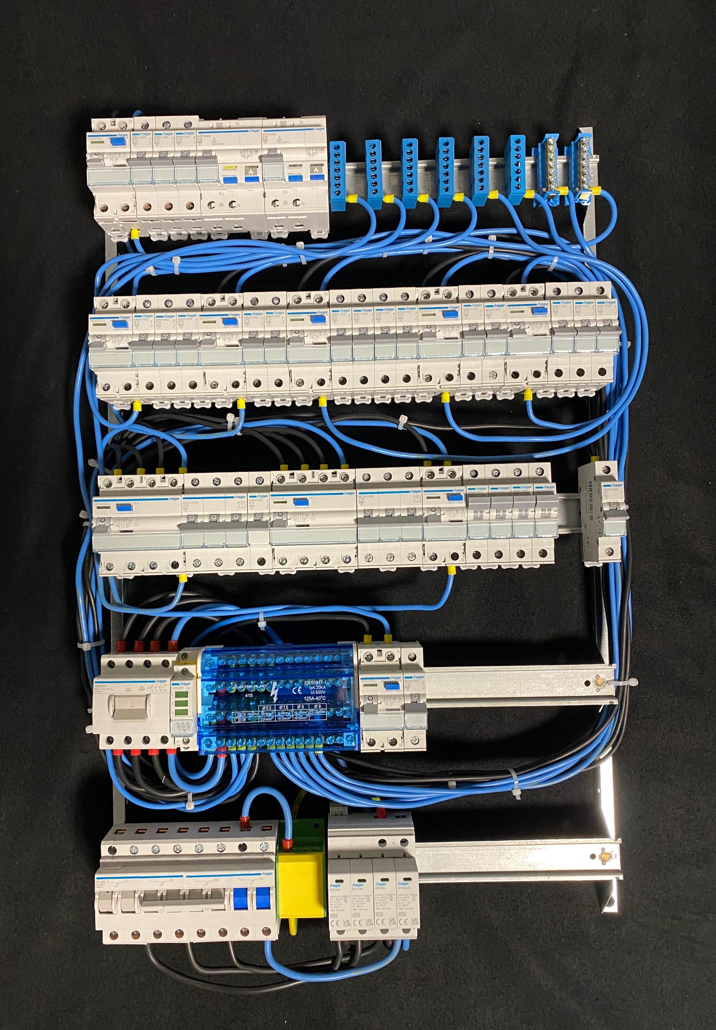 Rozdzielnica elektryczna Hager z zabezpieczeniami, modułami, czarnymi i niebieskimi przewodami, zamontowana na szynie w szafie elektrycznej na czarnym tle.
