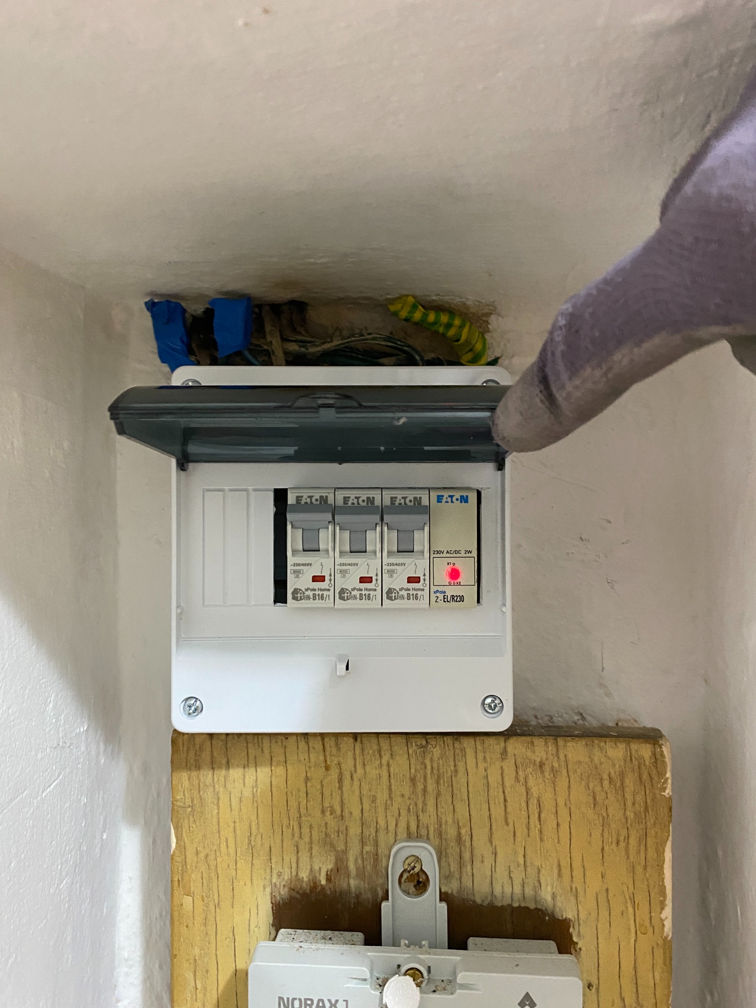 Otwarta skrzynka z bezpiecznikami Eaton na ścianie, z widocznymi kablami w ścianie i ręką w rękawicy wskazującą na wnętrze. Widoczny element instalacji elektrycznej.