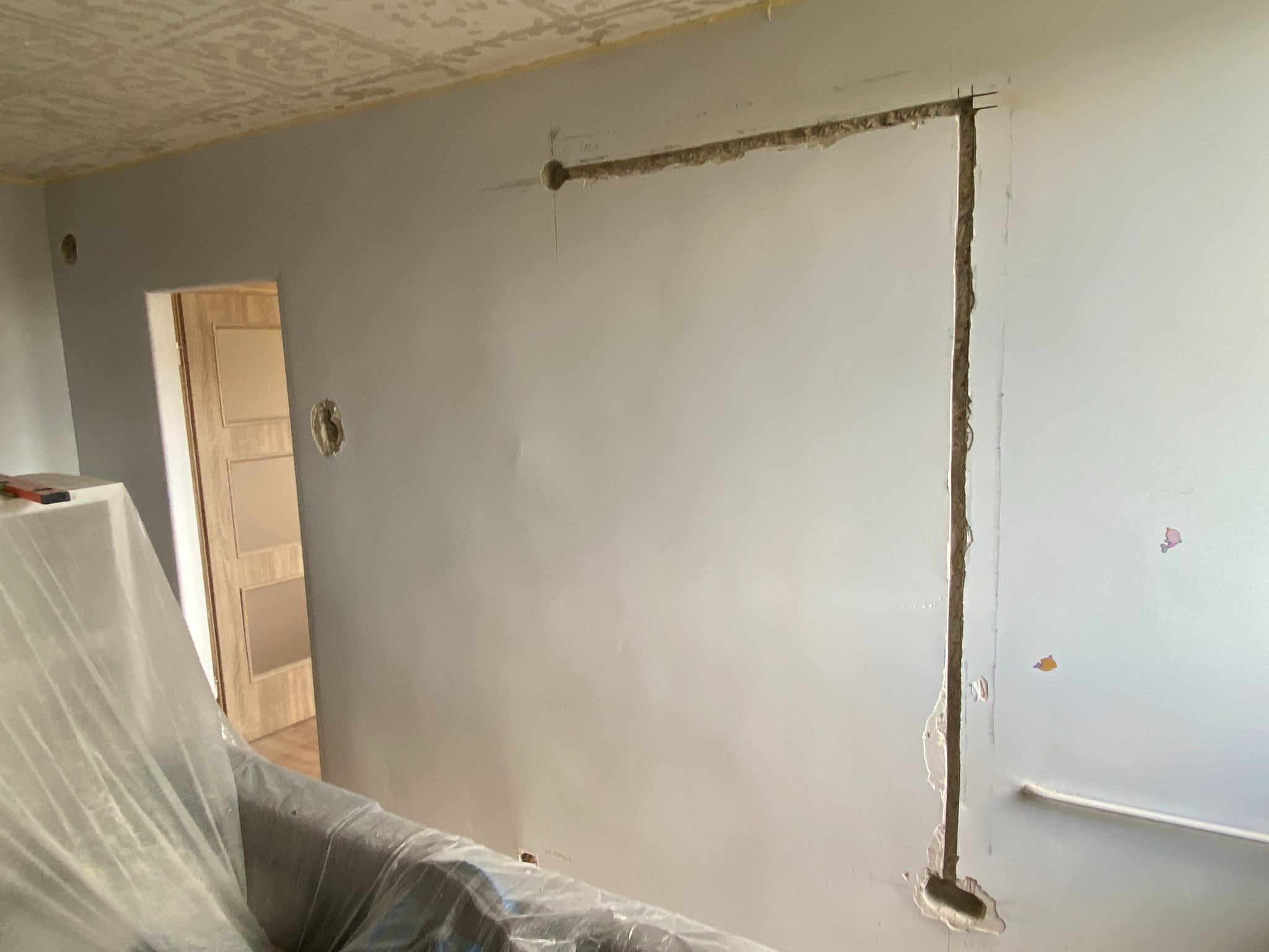 Ściana z wyżłobionymi bruzdami pod kable elektryczne, widoczne przygotowania do instalacji. Częściowo zakryte meble folią ochronną.