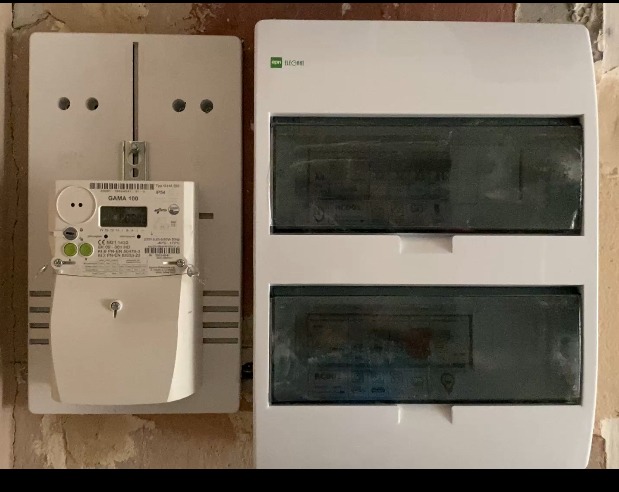 Nowy licznik energii elektrycznej GAMA 100 obok białej skrzynki bezpiecznikowej, zamontowane na ścianie z surową fakturą. Widoczne detale montażu i podłączenia.