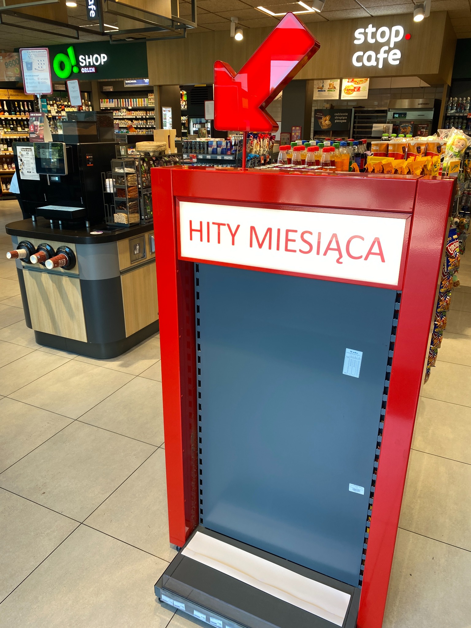 Półka sklepowa z napisem 'Hity Miesiąca' w czerwonej obudowie, z czerwoną strzałką na górze, w tle widoczne regały z produktami i kawiarnia Stop Cafe.