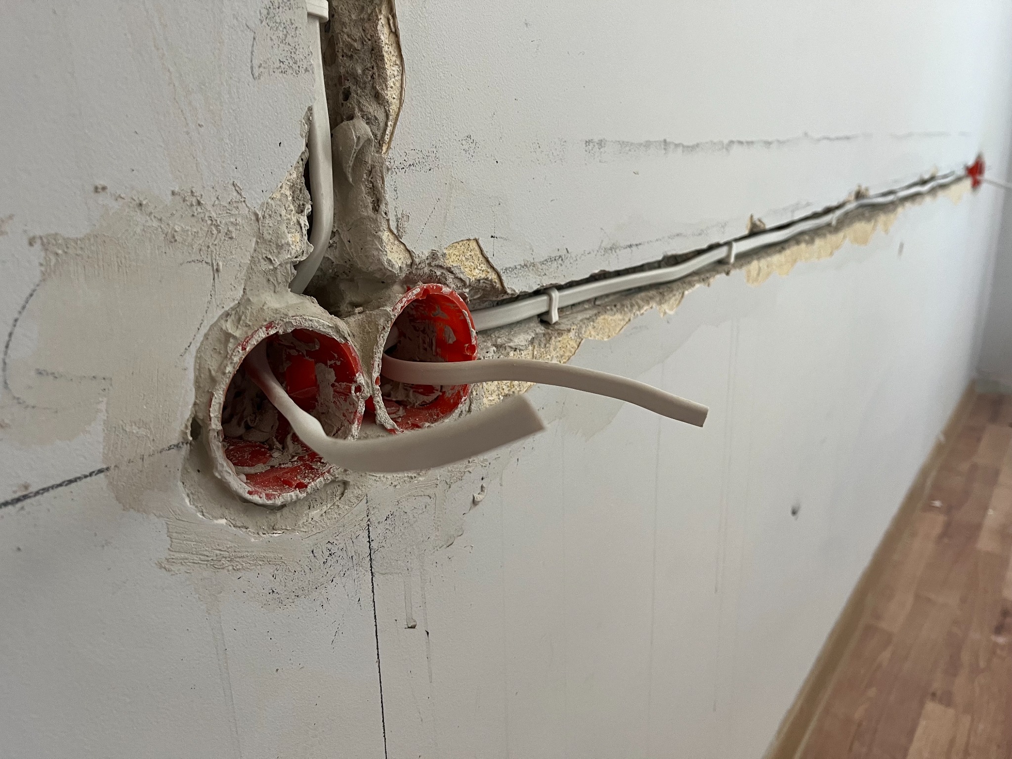 Ściana z dwoma czerwonymi puszkami instalacyjnymi i przewodami. Wykute bruzdy w ścianie z ułożonymi kablami. Widoczne ślady po gipsowaniu.