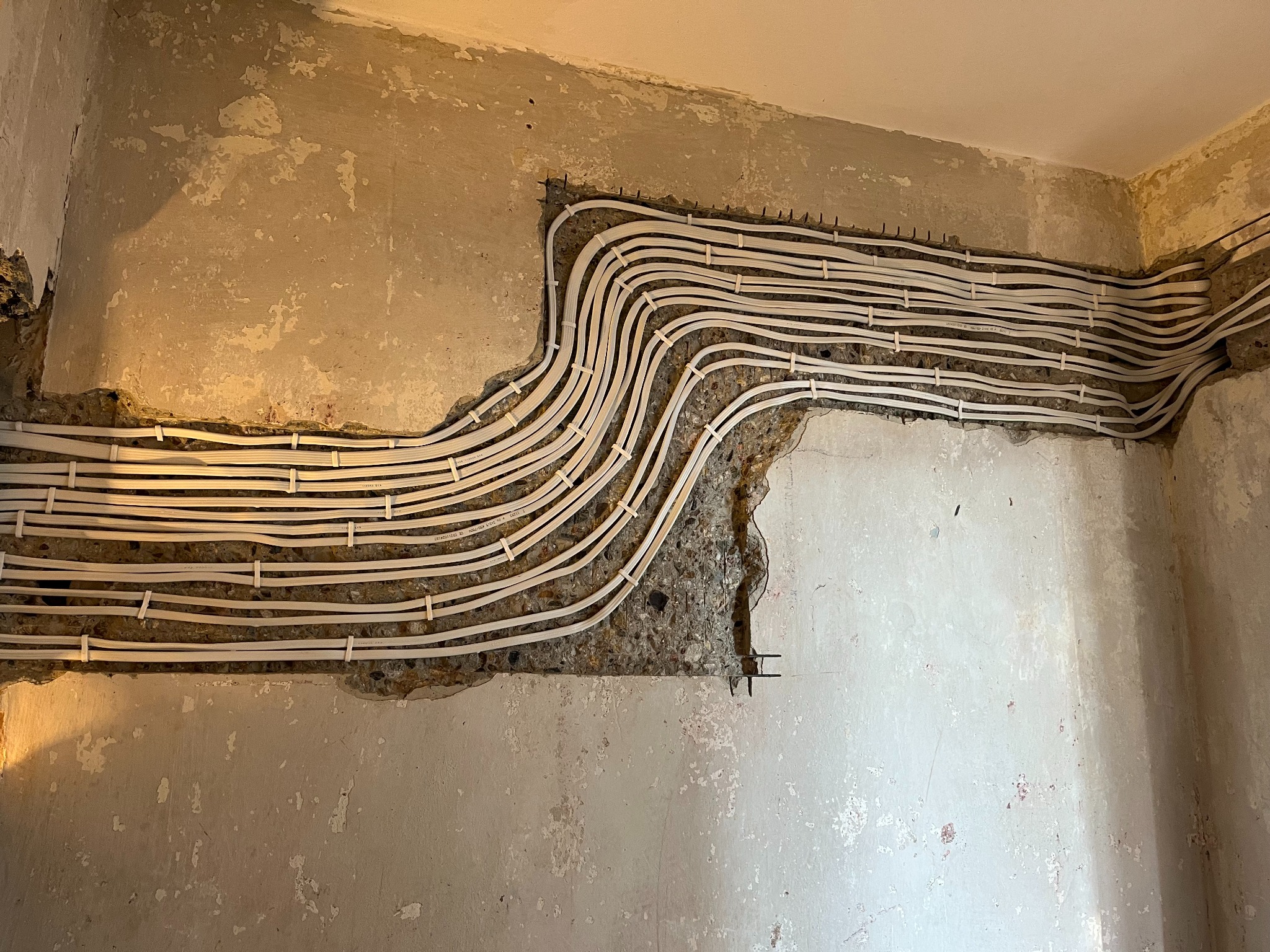 Białe przewody elektryczne ułożone w bruzdach ściany z surowego betonu, zabezpieczone uchwytami. Widok na fragment instalacji przed tynkowaniem w rogu pomieszczenia.