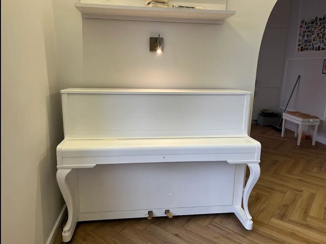 Białe pianino w jasnym wnętrzu z parkietem w jodełkę, oświetlone kinkietem. Widoczna półka nad instrumentem i fragment drugiego pomieszczenia w tle.