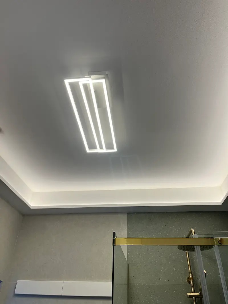 Nowoczesna lampa sufitowa LED w kształcie prostokąta, oświetlająca łazienkę z kabiną prysznicową i podświetlanym sufitem. Minimalistyczny design, jasne światło.