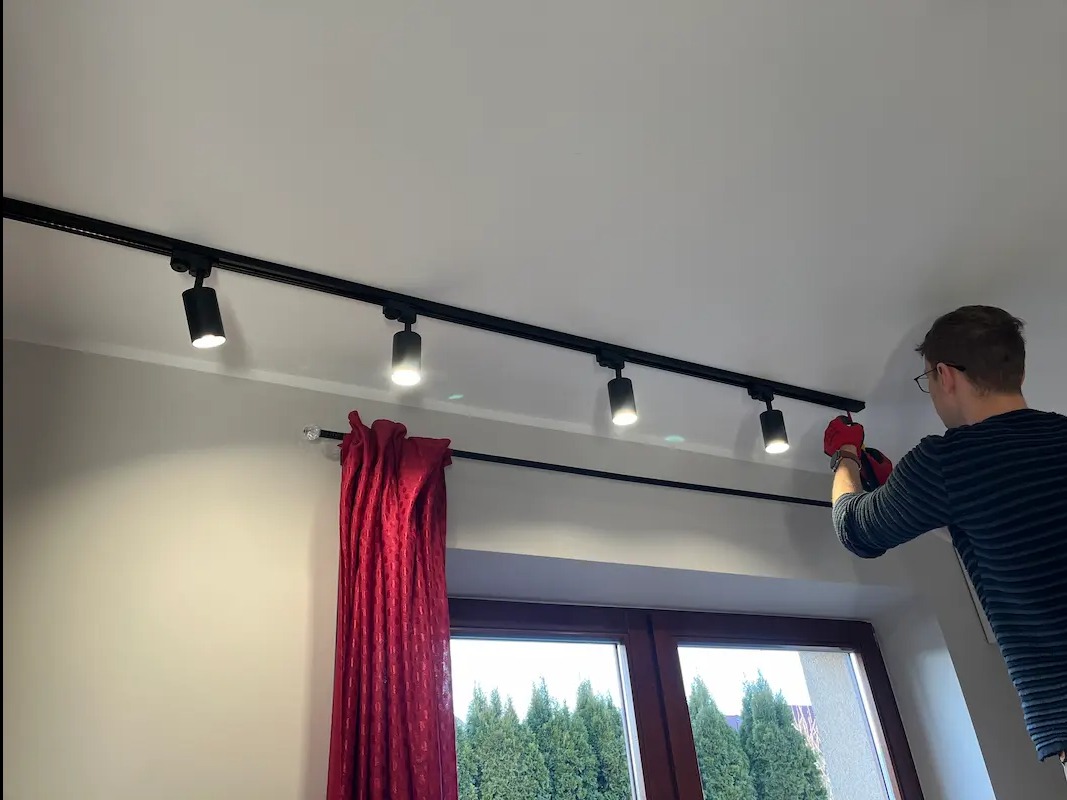 Montaż czarnej szyny z reflektorami sufitowymi w pokoju z czerwonymi zasłonami. Elektryk w czerwonych rękawicach instaluje oświetlenie.