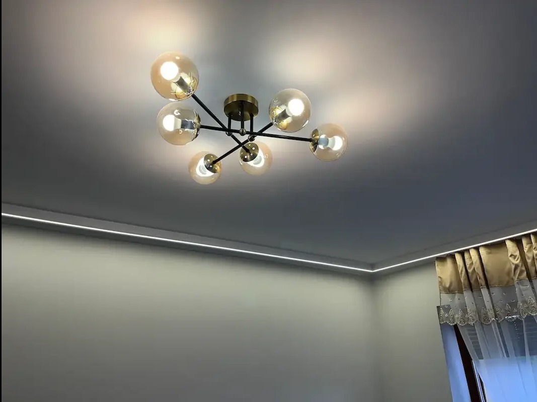 Sufit z nowoczesną lampą z kulistymi, bursztynowymi kloszami i listwą LED wzdłuż krawędzi. Widok z dołu, fragment zasłony w rogu.